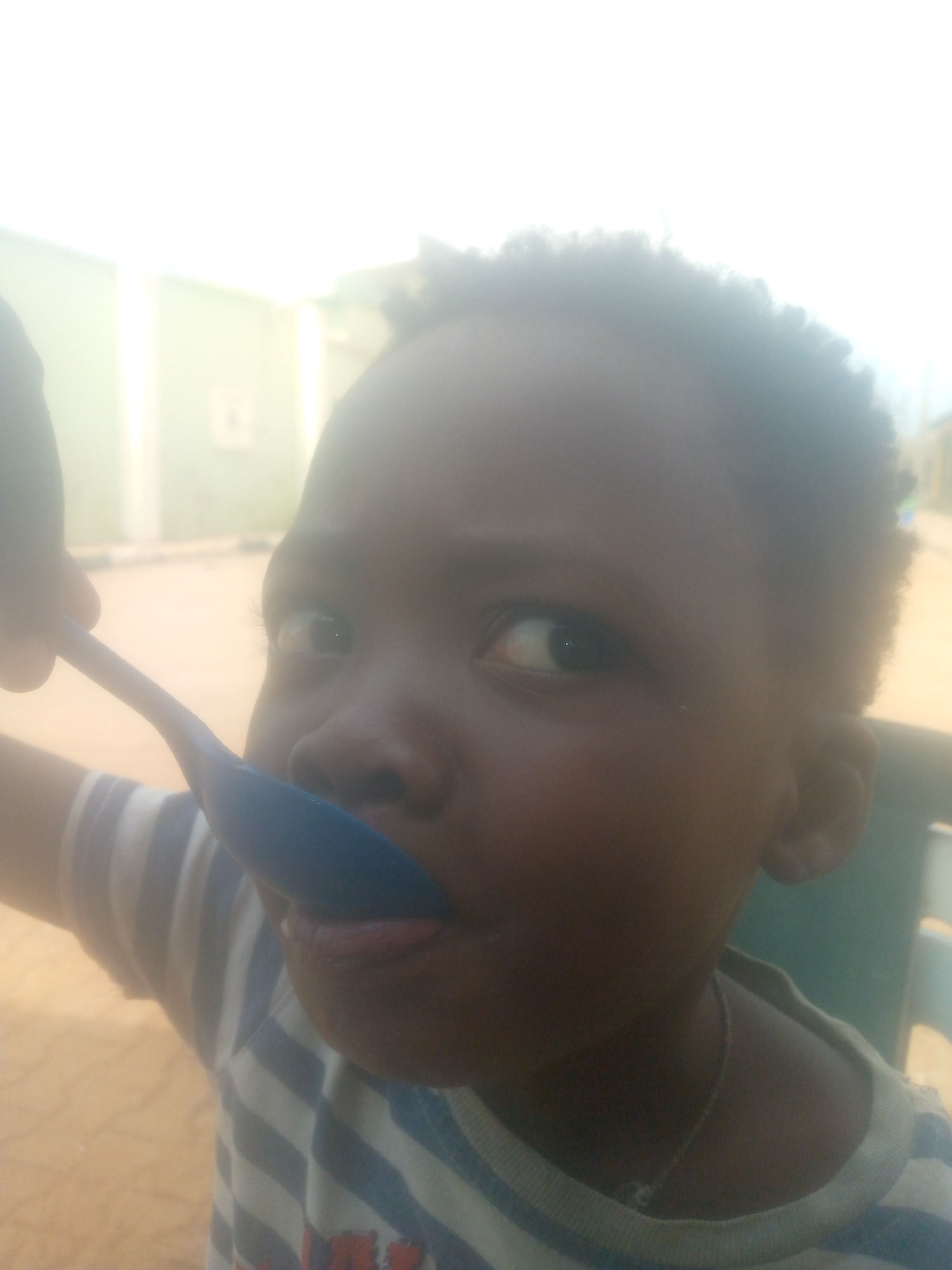 The image captures a close-up of a young child, likely a boy, with dark skin and short, dark, curly hair. The child's face and upper torso are visible, positioned slightly off-center and looking towards the right side of the frame. A blue spoon is at the child's mouth, with the tongue slightly extended, suggesting they are eating or about to eat from it. An adult hand, partially visible on the far left, holds the spoon.
The child is wearing a striped t-shirt with alternating white and dark blue or black horizontal stripes. A thin, silver-colored chain is visible around their neck. The background is softly out of focus and appears somewhat overexposed in brighter areas, particularly the top and left. Hints of a light green wall or structure are visible on the left, and a lighter, sandy-colored ground or pavement area fills the lower-middle background. A dark green object, possibly part of a chair, is partially visible on the right.
The bright lighting suggests the scene takes place during the daytime. The child's visible eye is wide open, giving an impression of curiosity or focused attention on the spoon. No text is visible in the image.