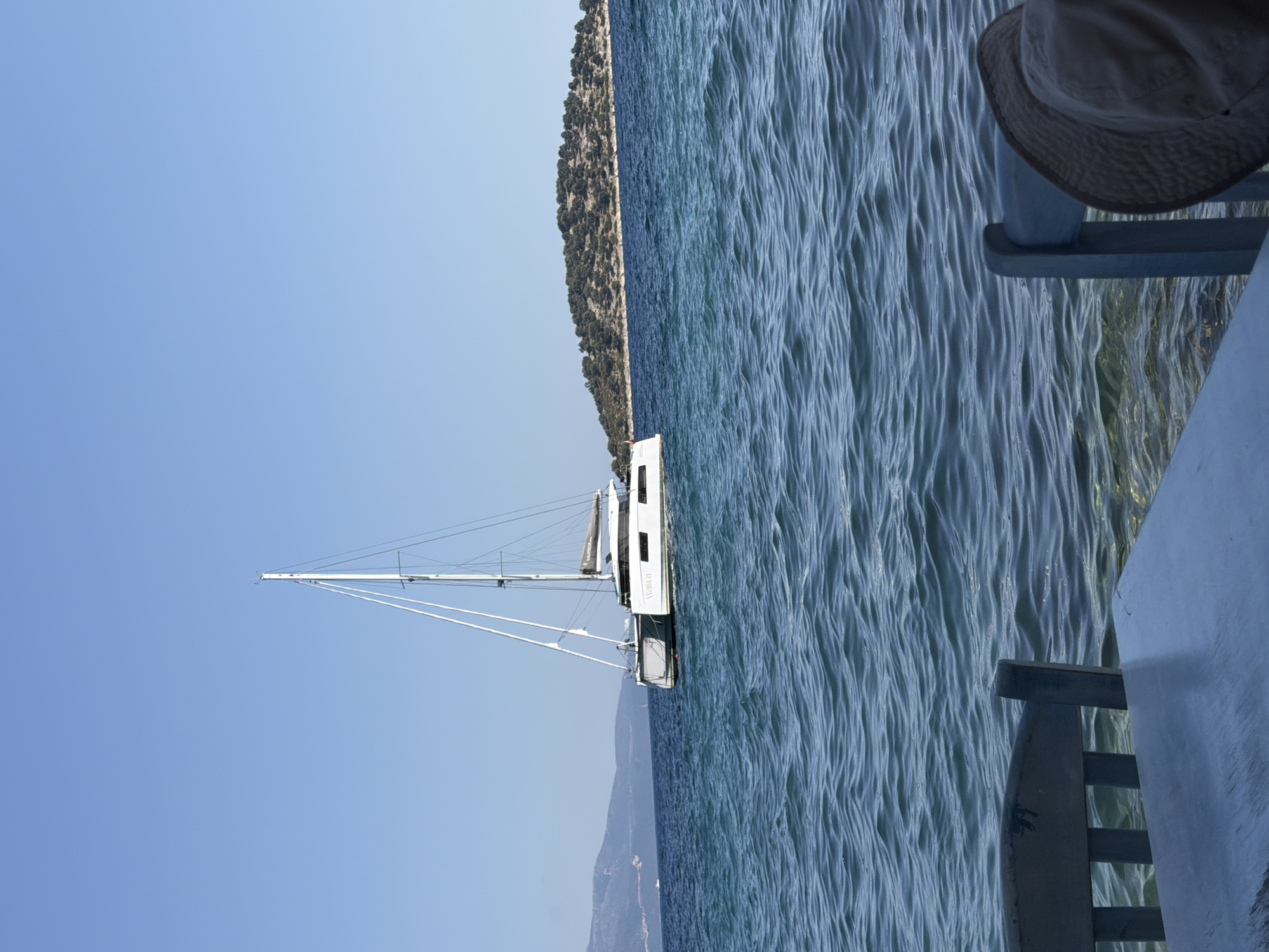 In Municipal Unit of Kalamos, Greece, a white catamaran sailboat with a tall mast is anchored in clear blue water under a cloudless sky. The catamaran is positioned in the middle distance, to the left of a small, tree-covered island. In the foreground, the edge of a table and two wooden chairs are visible, suggesting a seating area at the water's edge. A person wearing a sun hat is seated in one of the chairs. The water is calm with gentle ripples, and the background features a hazy mountain range.
