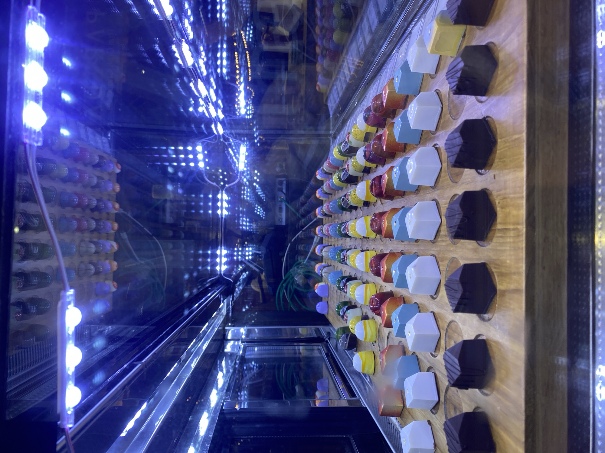 This media file captures a vibrant and artfully arranged display of colorful chocolates or candies within a glass display case, located in Choeng Thale, Thailand. The image is oriented vertically, showcasing rows of confectionery presented on a light wooden board with circular indentations for each piece.
The chocolates are diverse in shape and color. Some are dark, hexagonal, and feature a textured or fluted surface, resembling traditional dark chocolate bonbons. Interspersed among these are lighter-colored pieces, mostly square or rectangular prisms, boasting a smooth, glossy finish. These come in an array of bright hues, including white, light blue, yellow, orange, and reddish-brown. Many of the lighter candies are adorned with a delicate, irregular fleck of gold, likely gold leaf, which adds a luxurious detail. In the background, reflected within the glass, more rounded and multi-colored candies can be seen, hinting at the variety offered.
The display is dramatically illuminated by multiple strips of bright blue LED lights installed along the edges of the case. These lights create prominent reflections on the glass surfaces, giving the impression of an infinite tunnel of lights and duplicated rows of chocolates extending into the background. The intense blue light casts a cool, almost ethereal glow over the entire scene, making the vibrant colors of the chocolates pop. Visible wires connected to the LED strips are also present on the left side of the frame.
No people or direct activity are visible, suggesting a static product presentation. There is no visible text in the image. The scene conveys a sense of meticulous presentation and visual appeal, designed to attract customers to the confectionery on display.