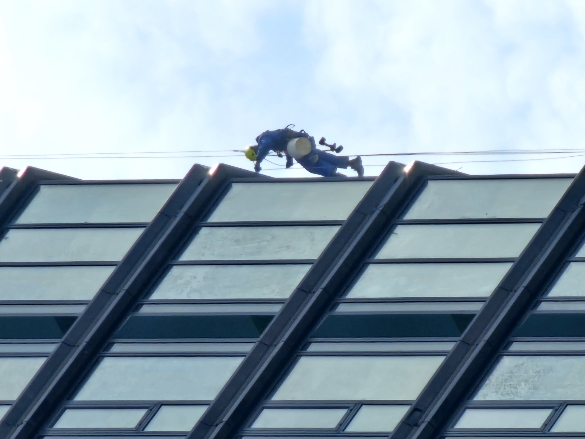 A window washer is visible on the slanted roof of a building. The worker is wearing a blue uniform and a yellow helmet and is equipped with a bucket and safety harness. The building has a modern design with diagonal support beams and glass windows. The sky is visible in the background. The location is Moscow, Russia.
