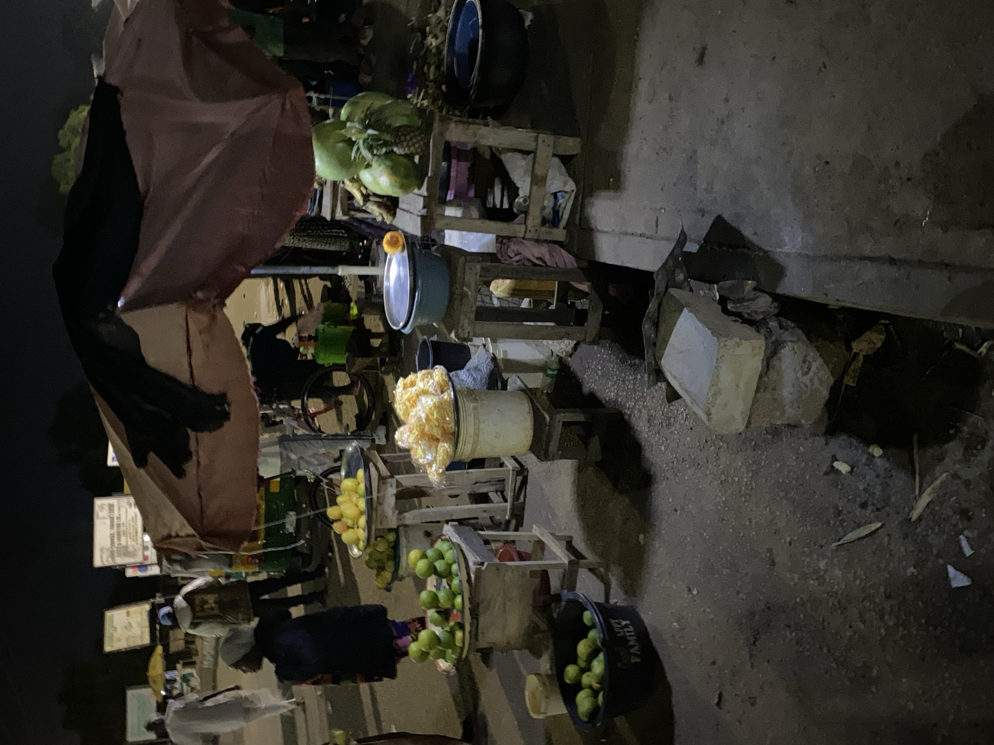 The image depicts a nighttime street scene in Bukuru, Nigeria, likely a market or roadside stall area. In the foreground, a dark plastic basin filled with green limes sits on the ground. To its right, a small wooden stand displays a metal tray with more green limes, some of which are stacked. Further back, another wooden stand holds a metal tray piled high with yellow fruits, possibly mangoes or papayas, and a large white bucket filled with what appears to be fried snacks or dried fruits. Adjacent to this, a larger wooden structure is laden with several watermelons and a pineapple. A large, dark blue basin is visible to the far right.
Overhead, a large, multi-colored umbrella, predominantly reddish-brown and black, provides shade. In the background, several people are visible. A person in white attire walks away from the camera on the left. Another individual in a patterned dress is partially visible behind the fruit stall. Further back, a person in a white hooded jacket and dark pants is seen near a green and yellow auto-rickshaw. Signs with text are mounted on poles in the distance, some illuminated. The ground is paved, with visible debris and shadows cast by the overhead lighting and structures.