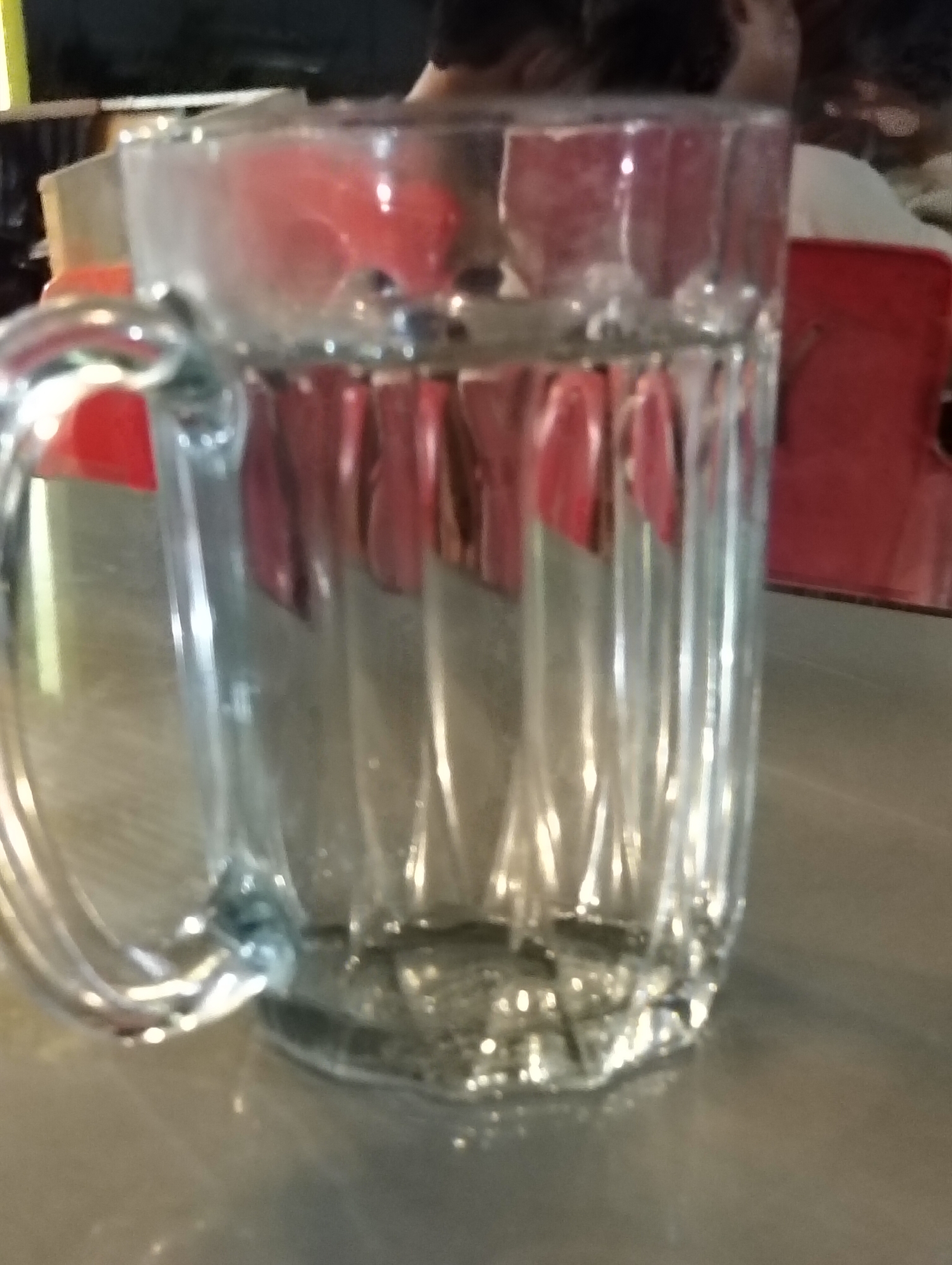 A clear glass mug with a handle, filled with a clear liquid, sits on a metallic surface. The mug is cylindrical with vertical ridges. Red objects are visible through the mug, reflecting on its surface. The background is dark and out of focus, with hints of red and white. The location is not discernible from the image.