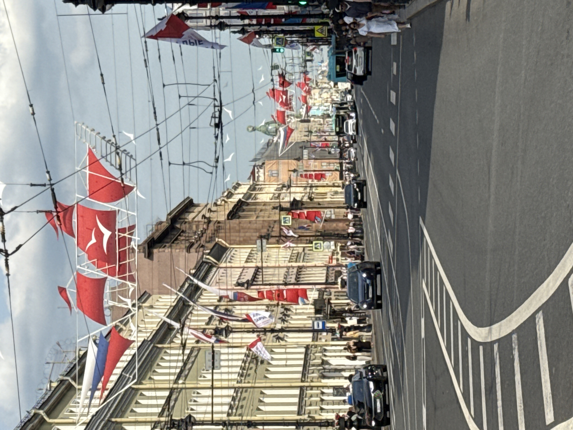 This image depicts a street scene in Saint Petersburg, Russia, likely during a daytime event. The perspective is from a slightly elevated viewpoint, looking down a long, wide street lined with buildings. Red and white flags, some with white emblems, are strung across the street, suggesting a festival or celebratory occasion. White flags are also visible, as are flags of the Russian tricolor.
The buildings appear to be historic, with ornate facades and large windows. There are power lines and street lamps crisscrossing the sky. Cars, including at least two black sedans and a blue van, are driving down the street, indicating normal traffic flow alongside the event decorations. Pedestrians are visible on the sidewalks, some appearing to be watching the event. The sky is partly cloudy, suggesting it could be a pleasant but not intensely sunny day.
Some visible text on a white flag appears to be in Cyrillic script, though it is partially obscured and difficult to read clearly. The overall impression is of a vibrant urban environment during a special event. The flags and decorations create a festive atmosphere within the historic setting of Saint Petersburg.