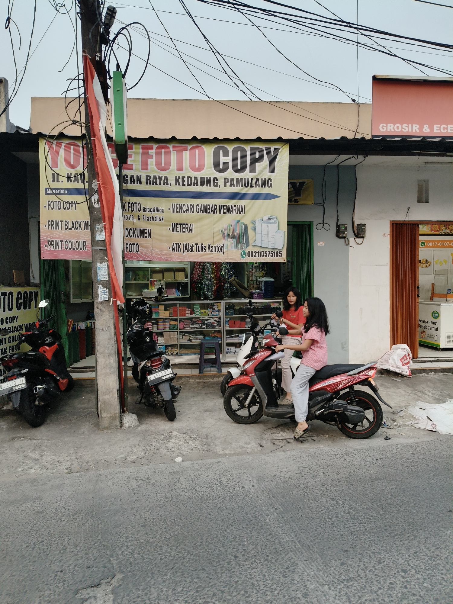 The image depicts a street scene in South Tangerang, Indonesia, in front of a photo copy shop. The shop has a large yellow banner advertising services like "FOTO COPY," "PRINT BLACK WH," and "PRINT COLOUR." Two young women are seated on a red and black motorcycle parked on the sidewalk. The woman in front is wearing a red and white checkered shirt and gray pants, while the woman behind her is wearing a pink t-shirt and light gray pants. They appear to be interacting or preparing to leave. Behind them, shelves are stocked with various goods, including stationery and possibly office supplies. Several motorcycles are parked along the sidewalk, indicating this is a common mode of transportation. The street is paved with concrete, and overhead power lines are visible against a pale sky. A prominent utility pole with the Indonesian flag attached stands to the left of the shop. The overall environment suggests a typical commercial street in an Indonesian city.