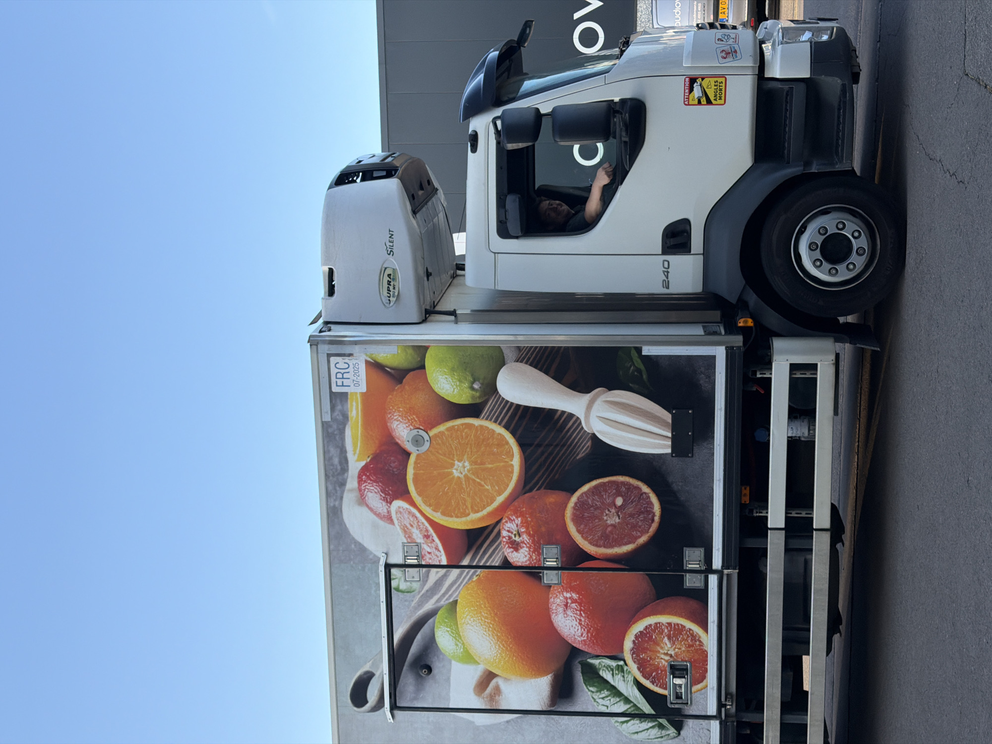 A white truck with a refrigerated unit is parked on an asphalt surface outdoors, under a clear blue sky. The side of the truck's cargo area features a large graphic of various fruits, including oranges, blood oranges, and limes, along with a wooden juicer. The truck has a driver visible inside the cab, smiling and looking towards the viewer. The driver is wearing a dark shirt. A warning sticker for "ANGLES MORTS" (blind spots) is present on the truck's exterior. In the background, a dark building with text is visible. The number "240" is marked on the side of the truck's cab. The scene appears to be in a commercial or industrial area, possibly Leudelange, Luxembourg, given the context provided.