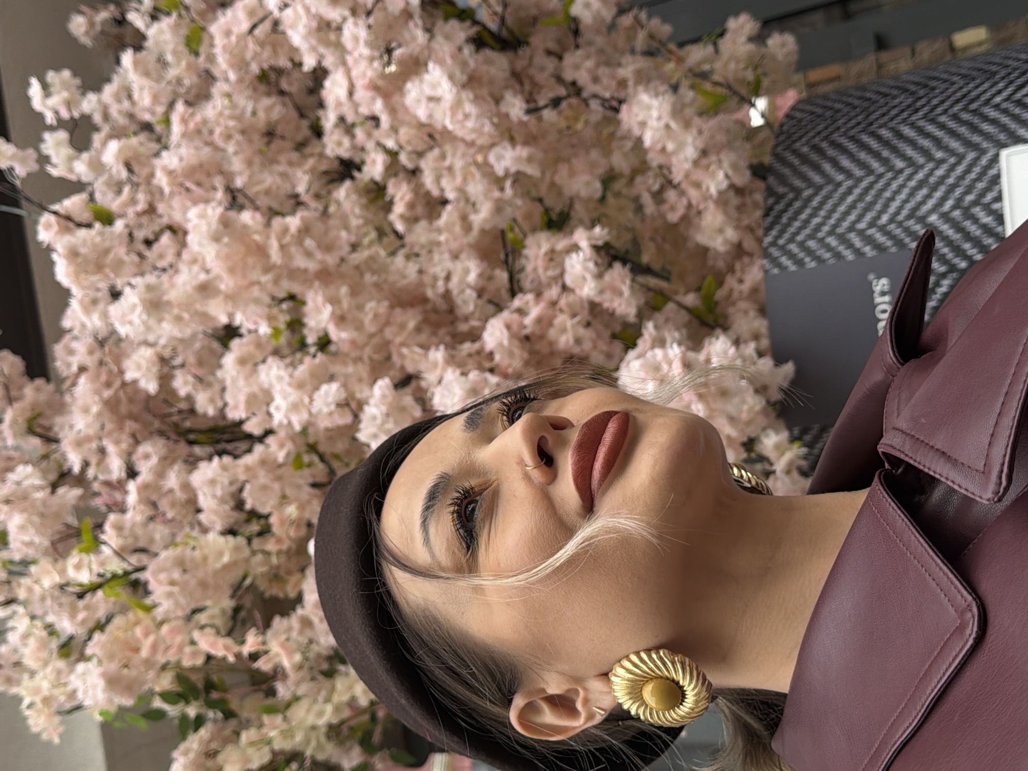 A close-up portrait captures a woman positioned to the right of the frame, looking upwards and slightly away from the camera with a serene expression, likely taken in Krasnoyarsk, Russia. She is set against a soft, blurred background dominated by numerous light pink artificial cherry blossoms, suggesting an indoor or decorated setting.
The woman has fair skin, prominent eyelashes, and defined eyebrows. She wears a brownish-red matte lipstick and a small gold nose ring. Her head is adorned with a dark brown beret, and large, textured gold earrings with a central button-like detail are visible on her earlobes. She is wearing a maroon-colored leather jacket with a structured collar.
In the upper right background, behind her shoulder, a blurred herringbone pattern in dark grey and white is visible, which could be part of furniture or a wall covering. Partial text "sors" is also faintly discernible on a dark surface in this area, likely part of a word or brand name. The lighting is soft and even, highlighting her features and the delicate floral backdrop.