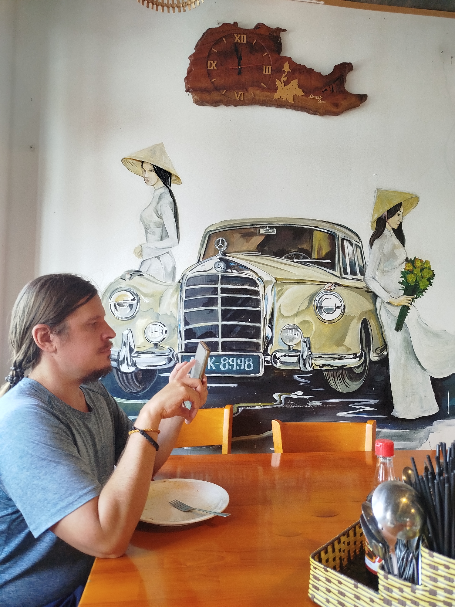 A medium shot captures a man with long hair tied in a braid, wearing a gray t-shirt, seated at a wooden table, and holding a smartphone. He appears to be engrossed in the phone. On the table is a half-eaten meal on a white plate with a fork. A condiments basket with spoons and chopsticks sits beside the plate, along with a bottle of chili sauce. Behind the man is a wall with a large mural depicting a vintage Mercedes-Benz car flanked by two women in traditional Vietnamese attire. Above the mural is a wooden clock shaped like a map. The location is Da Lat, Vietnam, as indicated by the text on the clock, "Souvenir Da Lat".