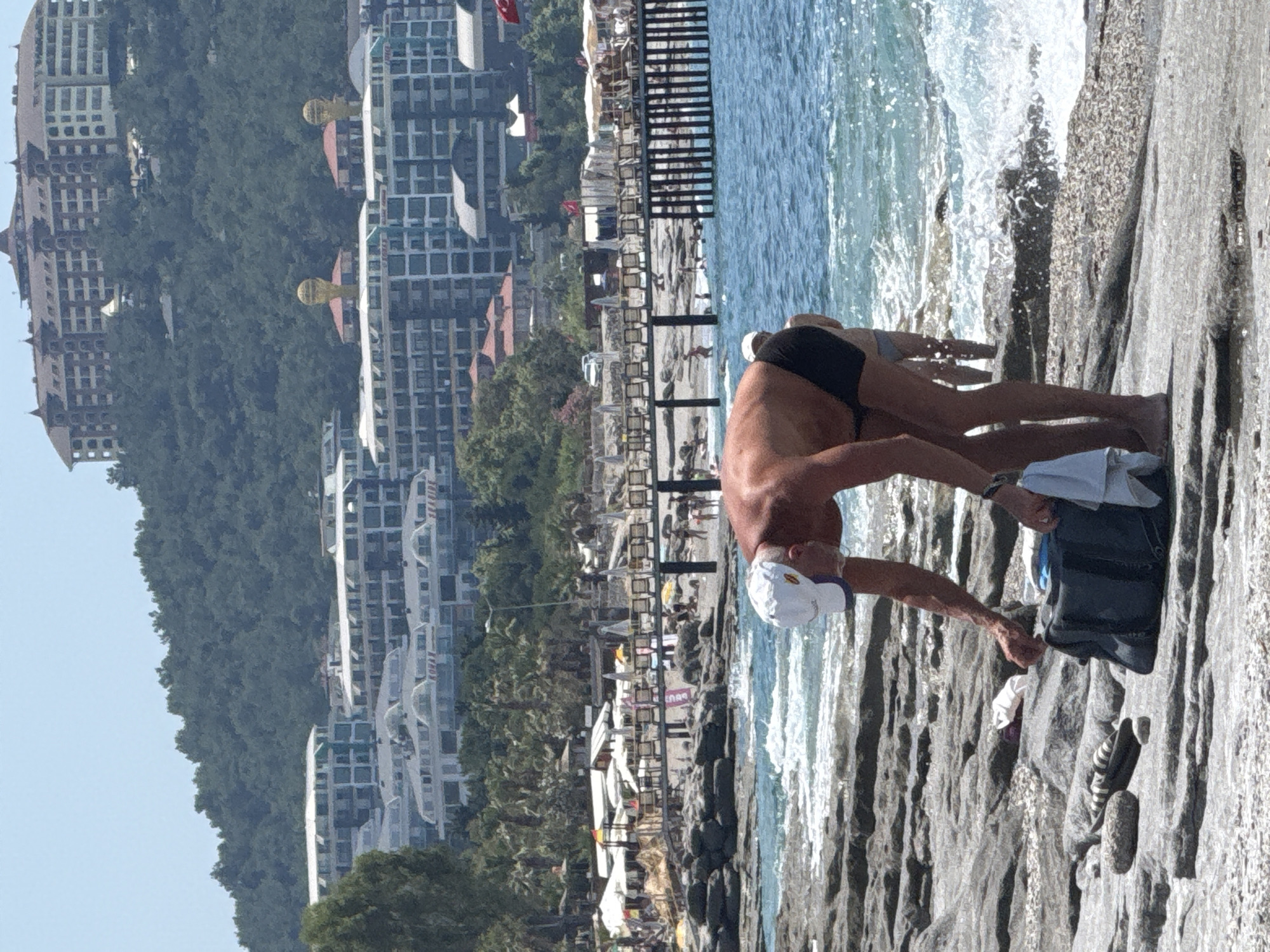 The image presents a sunlit coastal landscape in Alanya, Turkey. In the foreground, a fair-skinned man in black swim trunks and a white baseball cap is bent over a black duffel bag on a grey, rocky shoreline, appearing to retrieve or store an item with his right hand. A white cloth hangs from the bag, and a pair of striped flip-flops and another light-colored garment lie on the rocks nearby. To his right, the sea features breaking waves with white foam. The lower legs of another person, wearing light-colored swimwear, are partially visible entering the water.
The midground transitions to a blue sea that meets a sandy beach area on the left, where numerous white sun umbrellas and sun loungers are arranged. A wooden pier, also furnished with loungers, extends into the water from this beach. Multiple individuals are visible on the beach, the pier, and in the water. Palm trees line the area behind the beach amenities.
The background consists of a forested hill under a clear, light blue sky. On the upper part of the hill, a large, multi-story building with a distinctive reddish-brown roof and multiple turrets is prominent. Below this, nestled among the trees, are several modern, multi-story buildings with white roofs and numerous windows. Two of these lower structures feature distinctive golden, globe-shaped decorative elements on their rooftops.