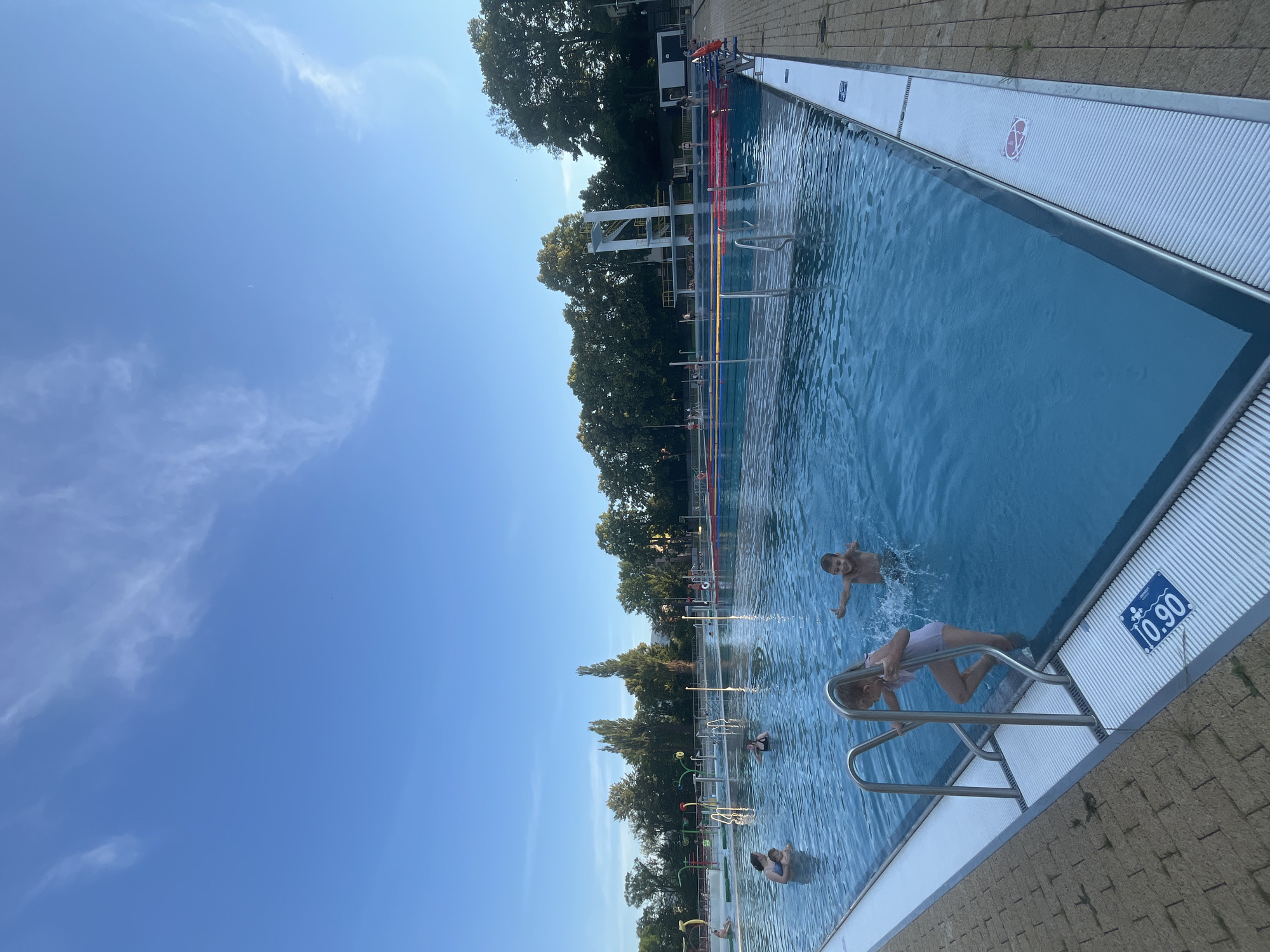 An outdoor swimming pool is shown on a clear day. Several people are visible in the water, including children playing and adults relaxing. The pool is divided into lanes with colorful ropes, and there is a diving platform in the background. Trees and buildings line the edge of the pool area. The water is a vibrant blue, and the sky is a clear, bright blue with a few wispy clouds. The overall impression is one of a fun and relaxing day at the pool.