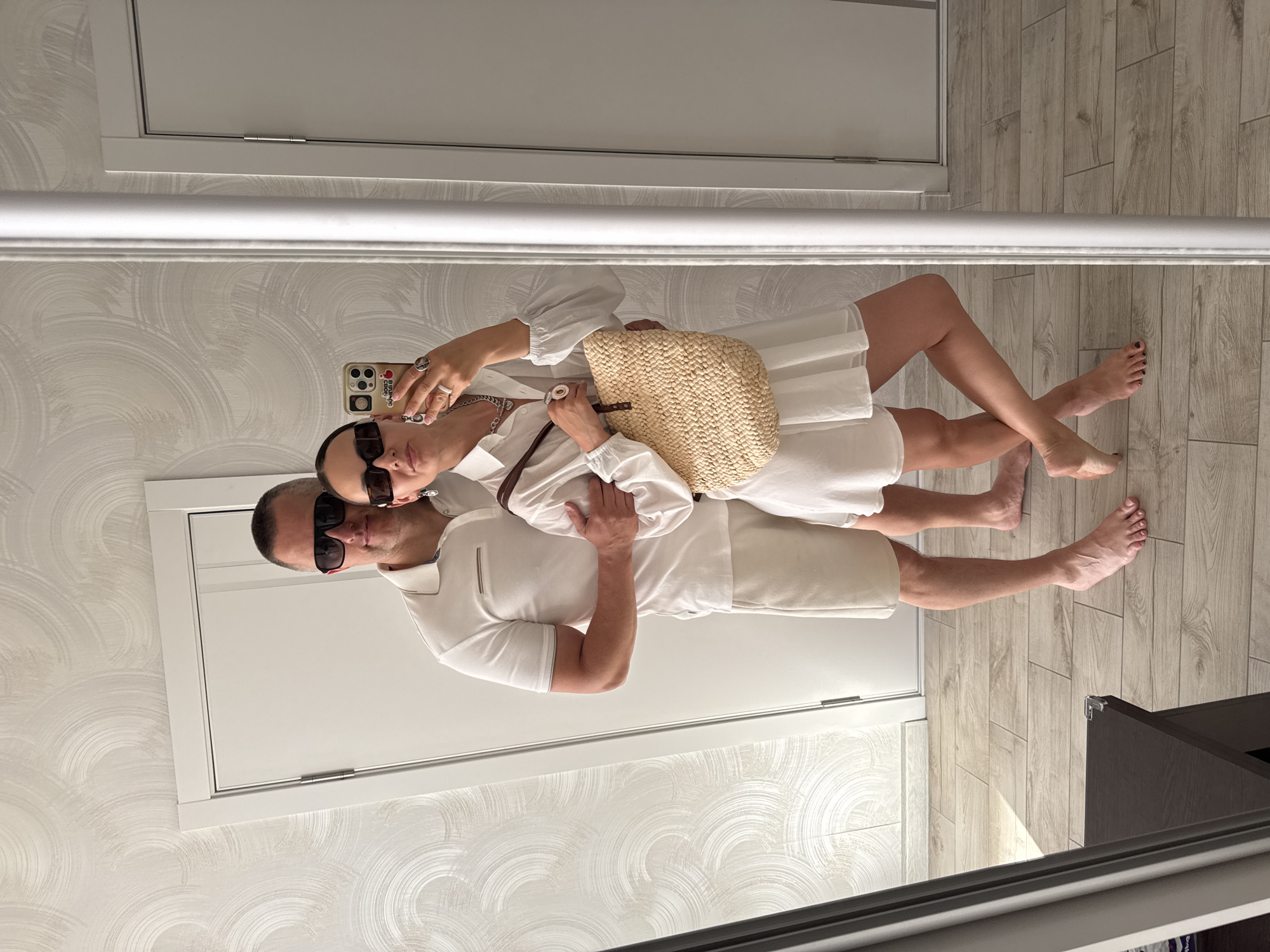 A man and a woman are taking a mirror selfie in an indoor location that could be a hotel room or apartment in Sochi, Russia. The woman is holding a mobile phone and a straw bag. The man has his arm around the woman. Both are wearing sunglasses. The woman is wearing a white dress and the man is wearing a white polo shirt and shorts. The floor is wood-grain tile. The walls have a textured wallpaper. There are two white doors visible in the background.