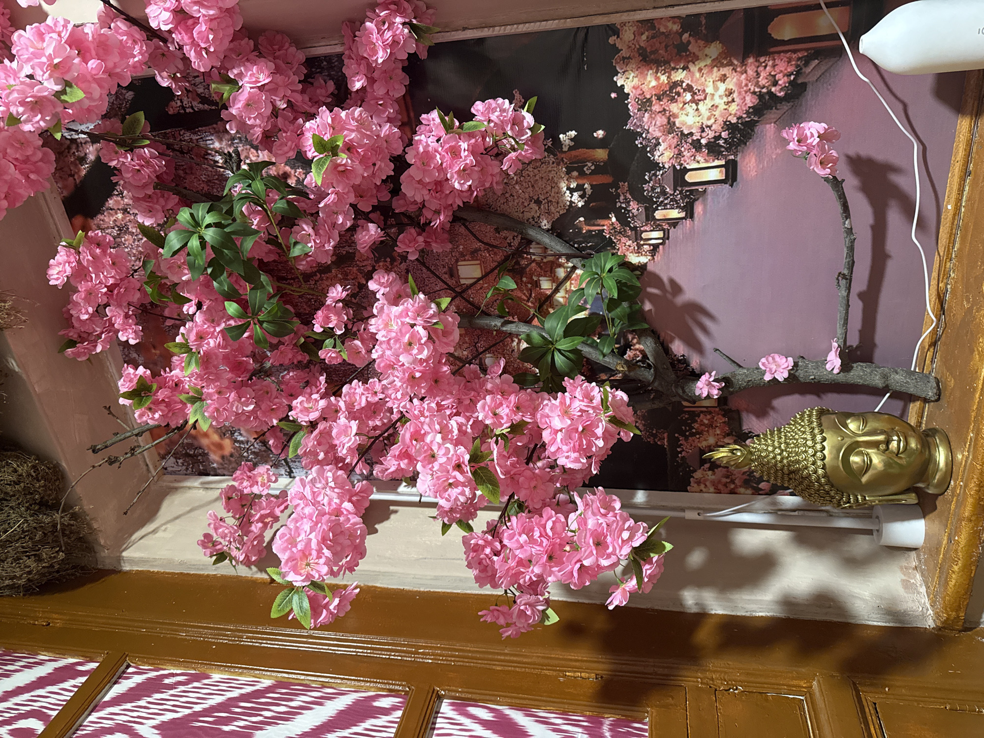 The image shows an interior space with a faux cherry blossom tree taking up a large portion of the view. The cherry blossoms are pink and full, with some green leaves visible. Behind the tree is a wall with a mural or wallpaper depicting an avenue of trees at night with illuminated streetlights.
On the right side of the image, a golden Buddha head is positioned near the wall. The lower portion of the image shows a wooden door or a part of a room divider with a reddish-purple and white patterned covering.