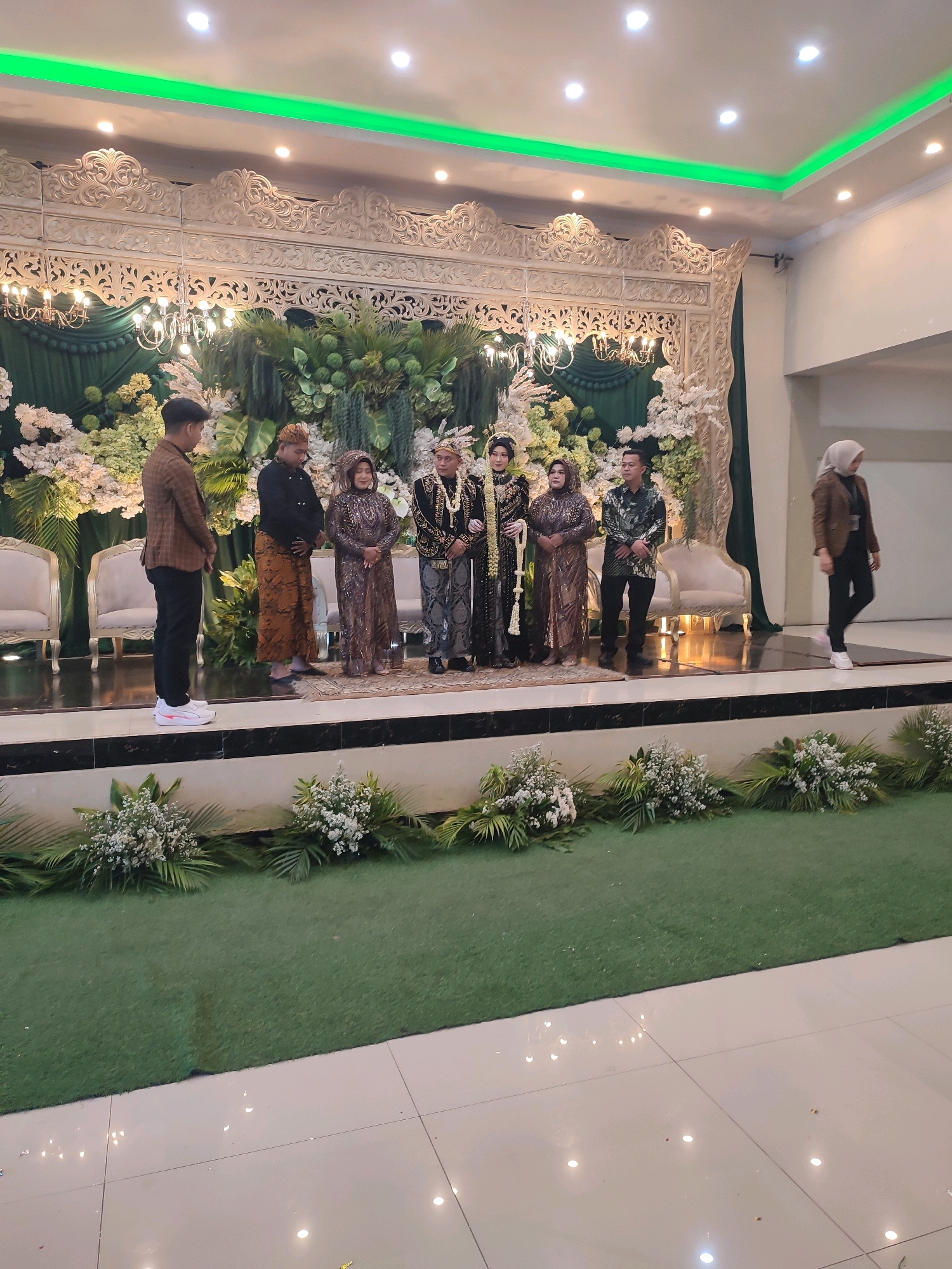 In Karangmalang, Indonesia, a wedding ceremony is taking place on an elevated stage adorned with ornate white carvings and lush green drapes. The stage is decorated with an abundance of white and green floral arrangements, creating a festive and elegant ambiance. In the foreground, artificial green grass covers the floor, bordered by further floral arrangements. A group of seven people, dressed in traditional Indonesian wedding attire, stand on the stage. The bride and groom, in the center, wear elaborate golden and black traditional garments. Flanking them are four individuals in similar ceremonial attire, and one person in a black and green patterned shirt and black pants. To the right of the stage, a woman in a hijab and a brown blazer is walking away from the scene. To the left of the stage, two white upholstered chairs are visible behind the floral arrangements. The ceiling features a green accent light and several white spotlights. The overall scene captures a traditional Indonesian wedding celebration in a well-decorated venue.