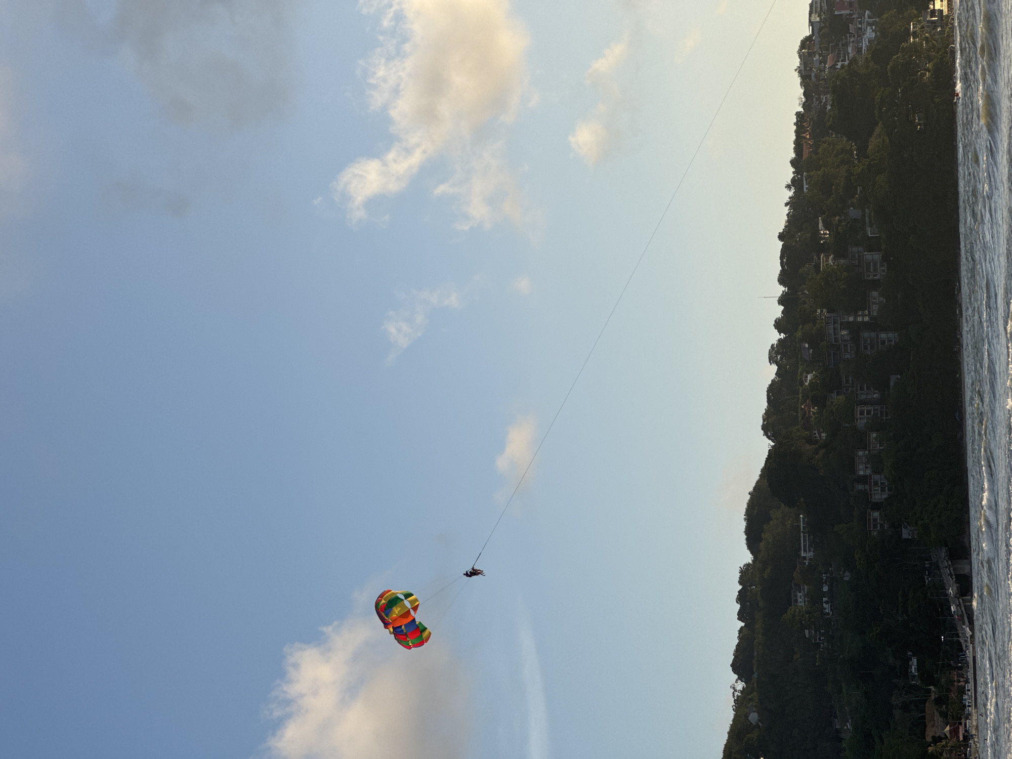 An aerial view shows a person parasailing over the ocean with a colorful parachute. In the background, a lush green hillside is dotted with buildings, suggesting a coastal town. The sky is a pale blue with scattered white clouds. The ocean in the foreground displays choppy waves, indicating a breezy day. The scene captures a moment of recreational activity against a scenic tropical backdrop.