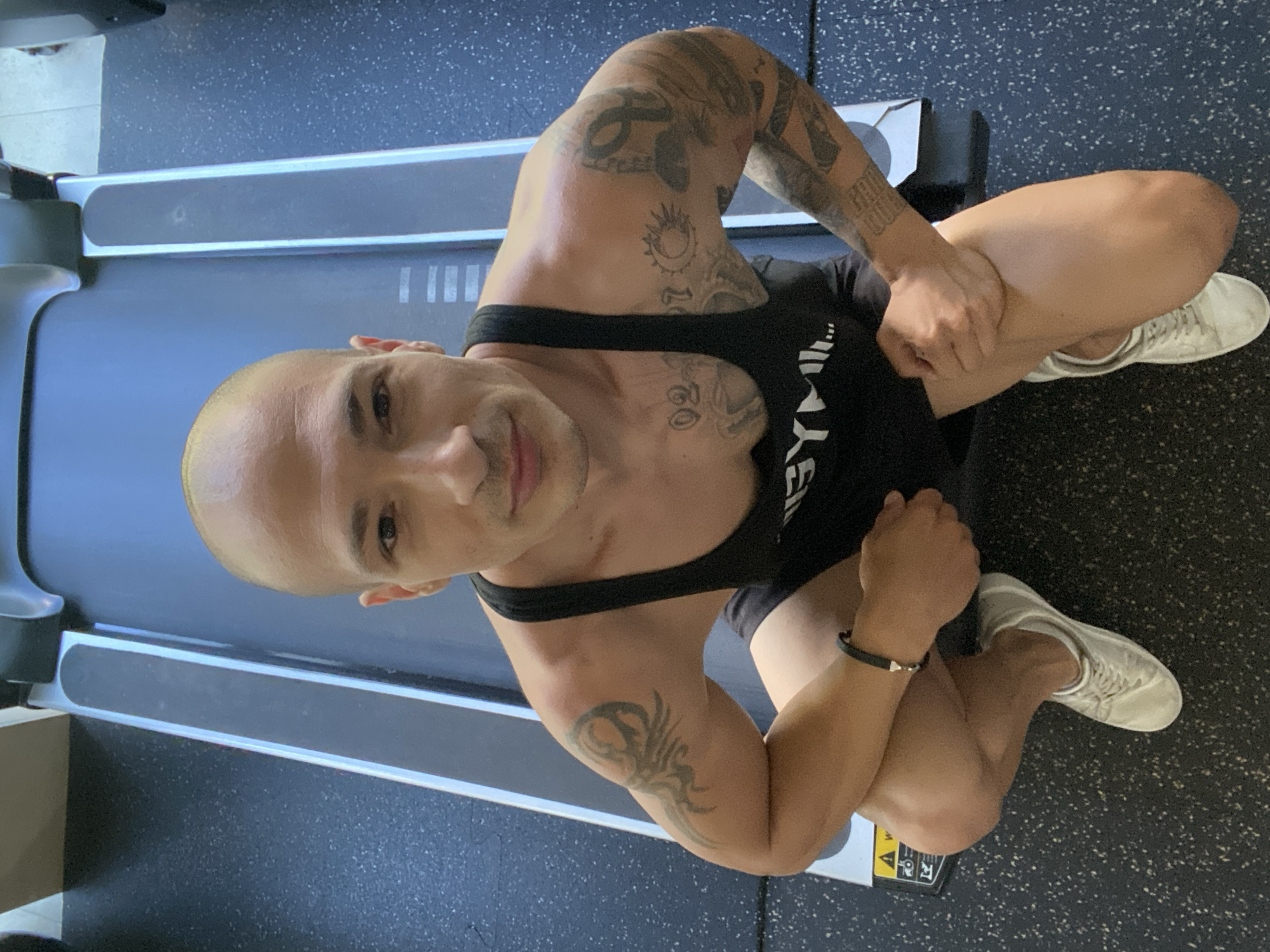 An adult male with a shaved head and visible tattoos on his arms and chest is captured in a high-angle, full-body shot. He is crouched down in front of a treadmill, which occupies the background. The man is wearing a black tank top with "GYML..." visible on it, black shorts, and white sneakers. He has a black bracelet on his left wrist. The floor is a dark, speckled material, typical of a gym environment. The image appears to have been taken indoors, likely in a gym or fitness center, possibly in the city of Pleven, Bulgaria, though this cannot be definitively confirmed from the visual evidence alone. The lighting is bright, suggesting overhead illumination. There are no other individuals or objects of significant note in the frame besides the man and the treadmill.