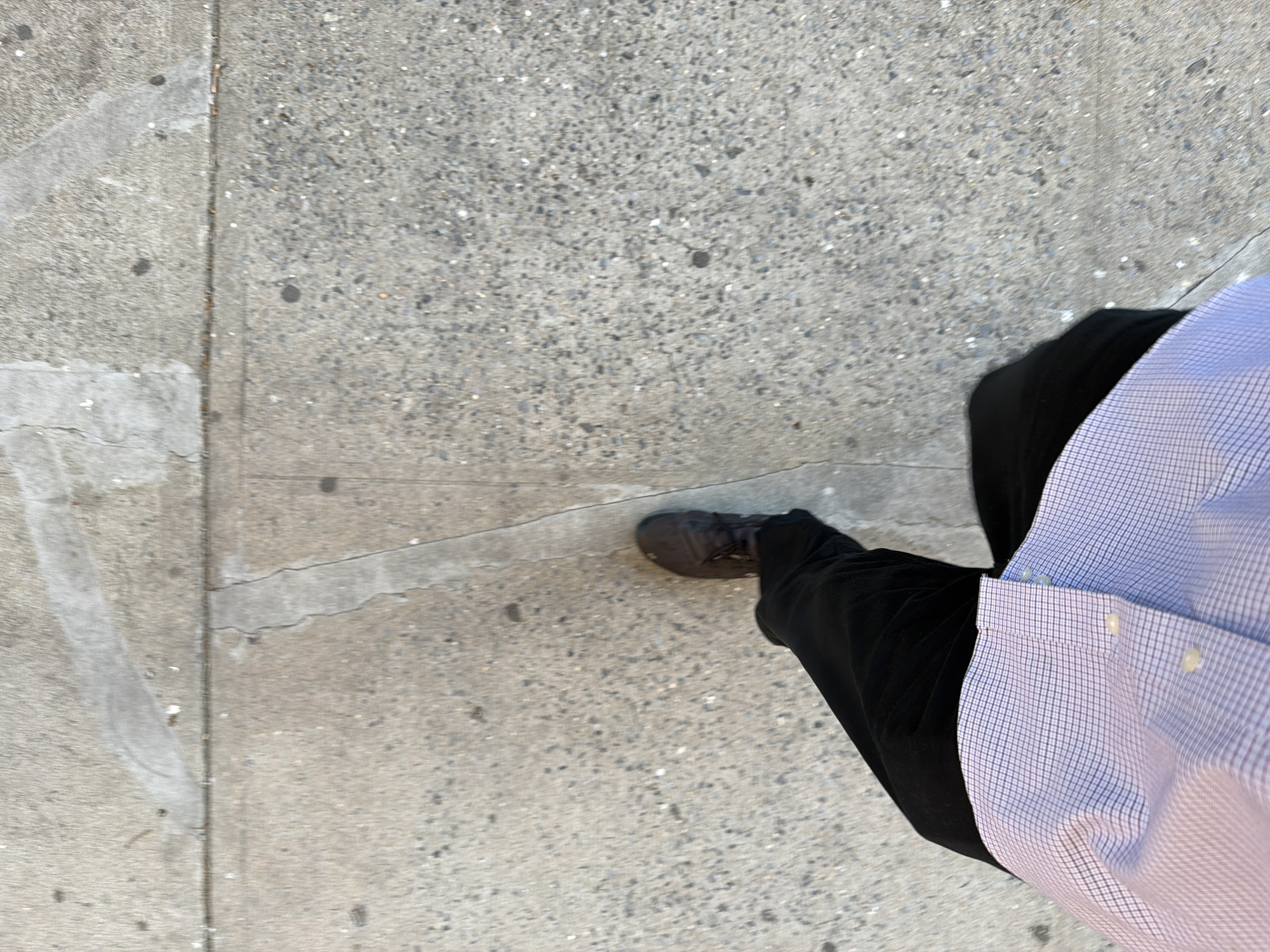 The image shows a first-person perspective of a person standing on a concrete sidewalk. The sidewalk appears to be in a city, likely New York, as evidenced by the typical urban pavement and the style of dress. The person is wearing black pants, a brown shoe, and a light blue and white checkered shirt. The focus is downward, showcasing the person's foot and leg on the sidewalk. The sidewalk has cracks and variations in texture and color.