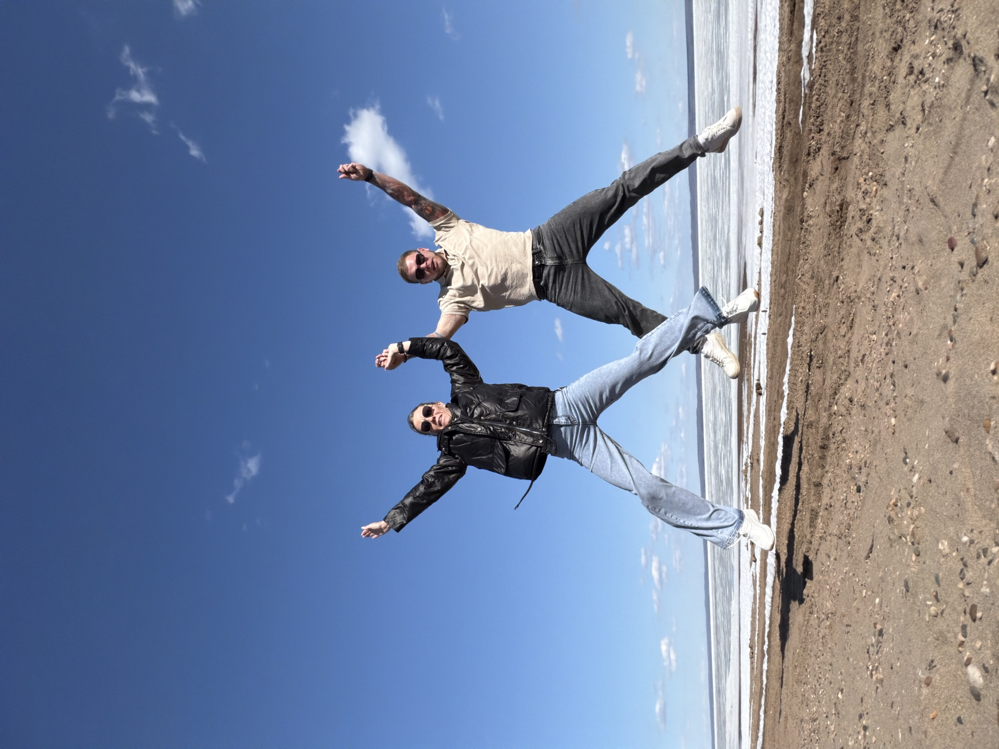 Two individuals are positioned in a way that they appear to be suspended in mid-air, with the sky visible behind them. The individual at the top wears a tan shirt and dark jeans, while the person below wears a black jacket and blue jeans. Both have their arms outstretched and legs spread apart.
The background includes a stretch of land and a body of water. Part of the water looks like a frozen area or ice.
Although the request mentioned the location, there is no definitive visible evidence in the image to independently verify it is in the city of Noviy, Russia. The image features details such as attire and an outdoor environment.