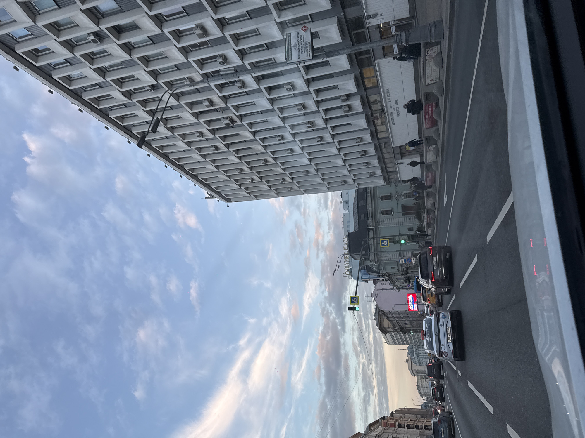 The image depicts a street scene in Moscow, Russia, with a large, modern building featuring a waffle-like patterned facade dominating the left side of the frame. A street stretches out into the distance, filled with cars moving in both directions. There is a clear sky with streaky clouds. The camera appears to be inside a vehicle, partially obstructed by the car window frame. The building is likely located on Prospekt Mira (Peace Avenue), as visible on a sign indicating a subway station and referencing V.I. Lenin. There is also a sign indicating a military commissariat of the city of Moscow. A sign displays “OLYMPIC PLAZA”. Traffic lights are visible, showing a green light. There are people present on the sidewalks. A car registration plate reads “C524MM 797”.