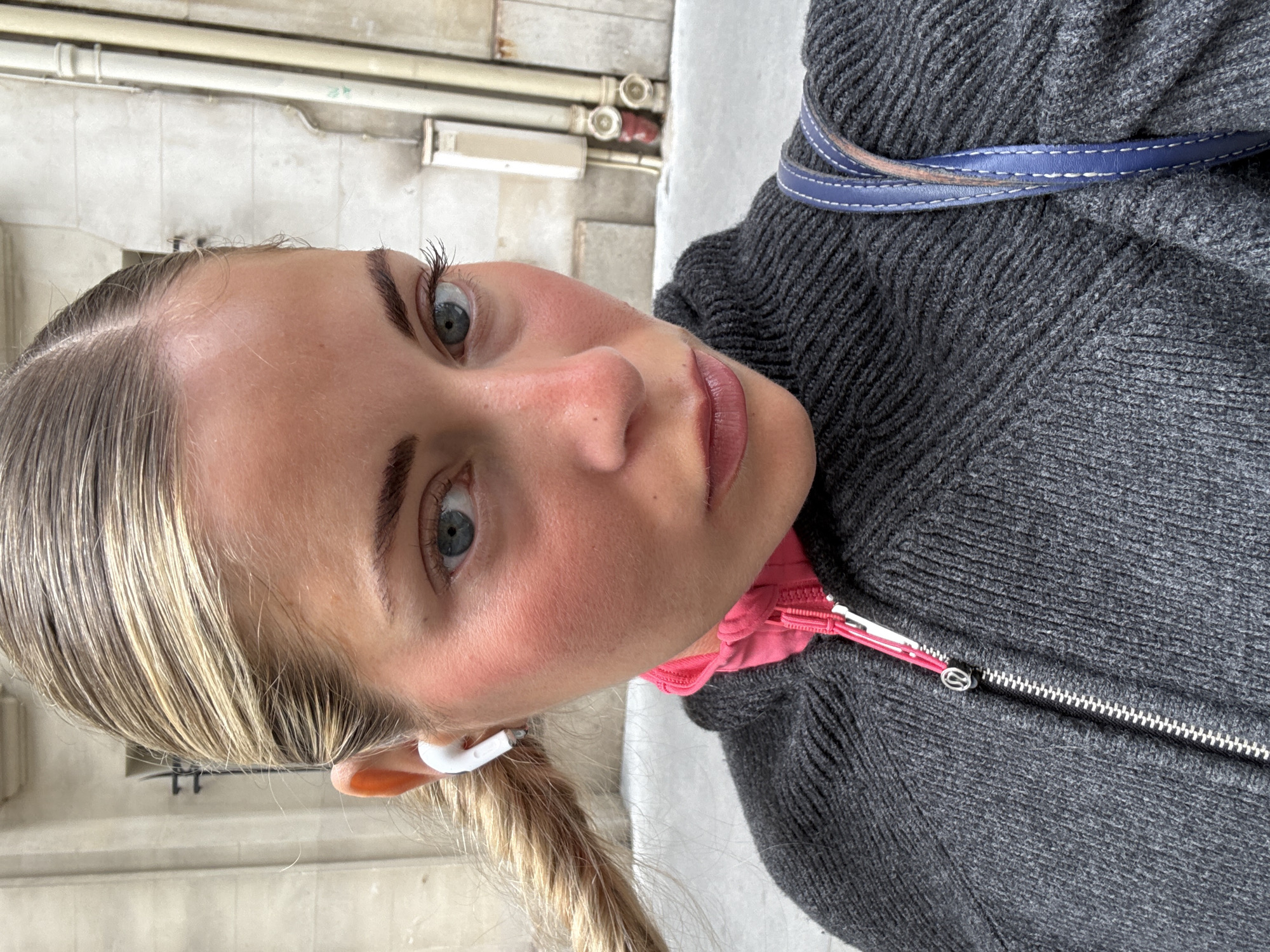 A selfie of a fair-skinned woman with blonde hair and blue eyes, wearing a gray sweater over a pink garment. She is wearing a wireless earbud in her left ear. The image is taken outdoors. The background suggests a Parisian setting with a weathered, light-colored building and pipes visible in the background.