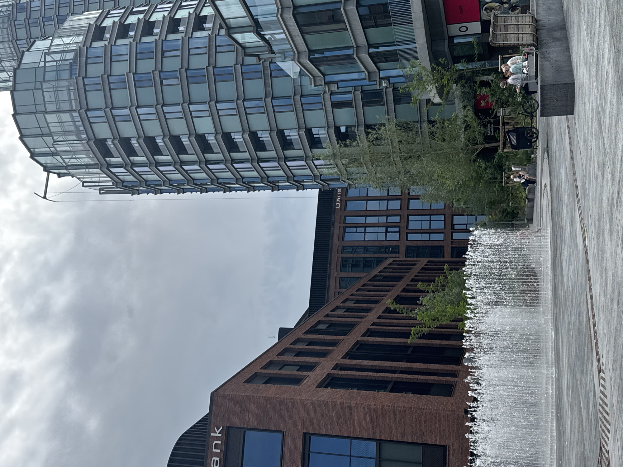 The image depicts a modern urban plaza in Copenhagen, Denmark, likely in the Nordhavn district, characterized by contemporary architecture and public amenities. In the foreground, a wide, shallow fountain sprays a dense curtain of water across the plaza's grey stone paving. To the left, a red brick building with dark-framed windows features signage that reads "bank" on its upper facade. Adjacent to this, and further into the midground, is another building with a dark facade and large windows. Dominating the right side of the frame is a tall, multi-story glass and concrete building with a distinctive curved facade and numerous balconies. Several trees and green spaces are visible between the buildings, adding a touch of nature to the urban landscape. In the midground, a group of people are seated on a long, concrete bench, some engaged in conversation, while a bicycle and a small cart are parked nearby. Another person is seated alone on a bench to the left. The sky is overcast, suggesting a cloudy day. The scene conveys a sense of public space designed for relaxation and modern city living.
