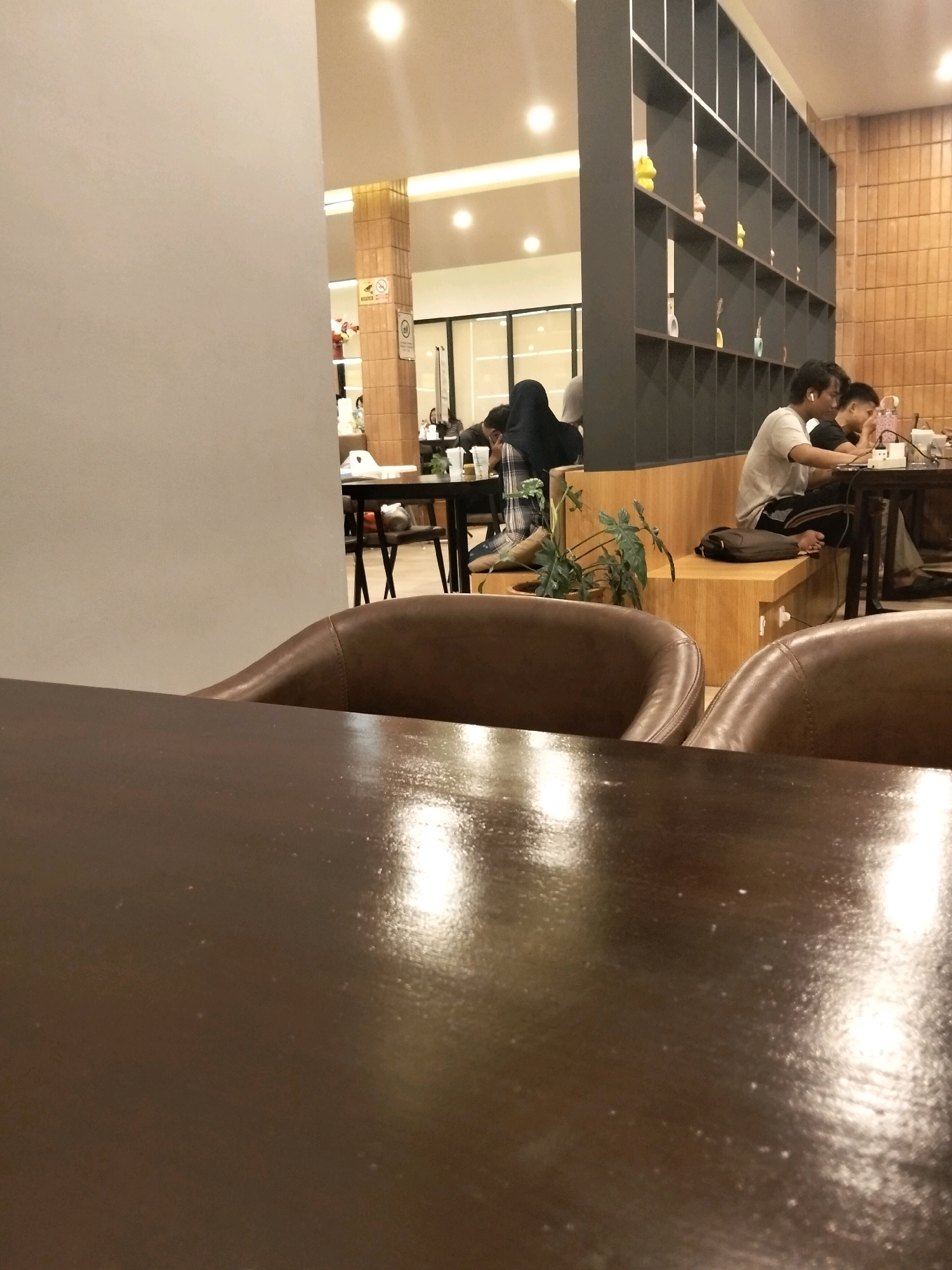 The image depicts the interior of a cafe, likely located in Arang Limbung, Indonesia. In the foreground, a dark, glossy table reflects overhead lights. Beyond the table, two plush, brown leather chairs are partially visible. In the mid-ground, a group of people are seated at tables. Two young men are at a table on the right, with one wearing a grey t-shirt and the other a black t-shirt, both appearing engaged with a laptop and cell phone. Another person, possibly a woman, is seated in the background on the left, wearing a dark hijab and looking down at a table. A large, dark grey bookshelf with multiple compartments filled with decorative items dominates the right side of the mid-ground, extending towards the background. The cafe environment includes wooden paneling on the walls and seating areas, and soft overhead lighting.