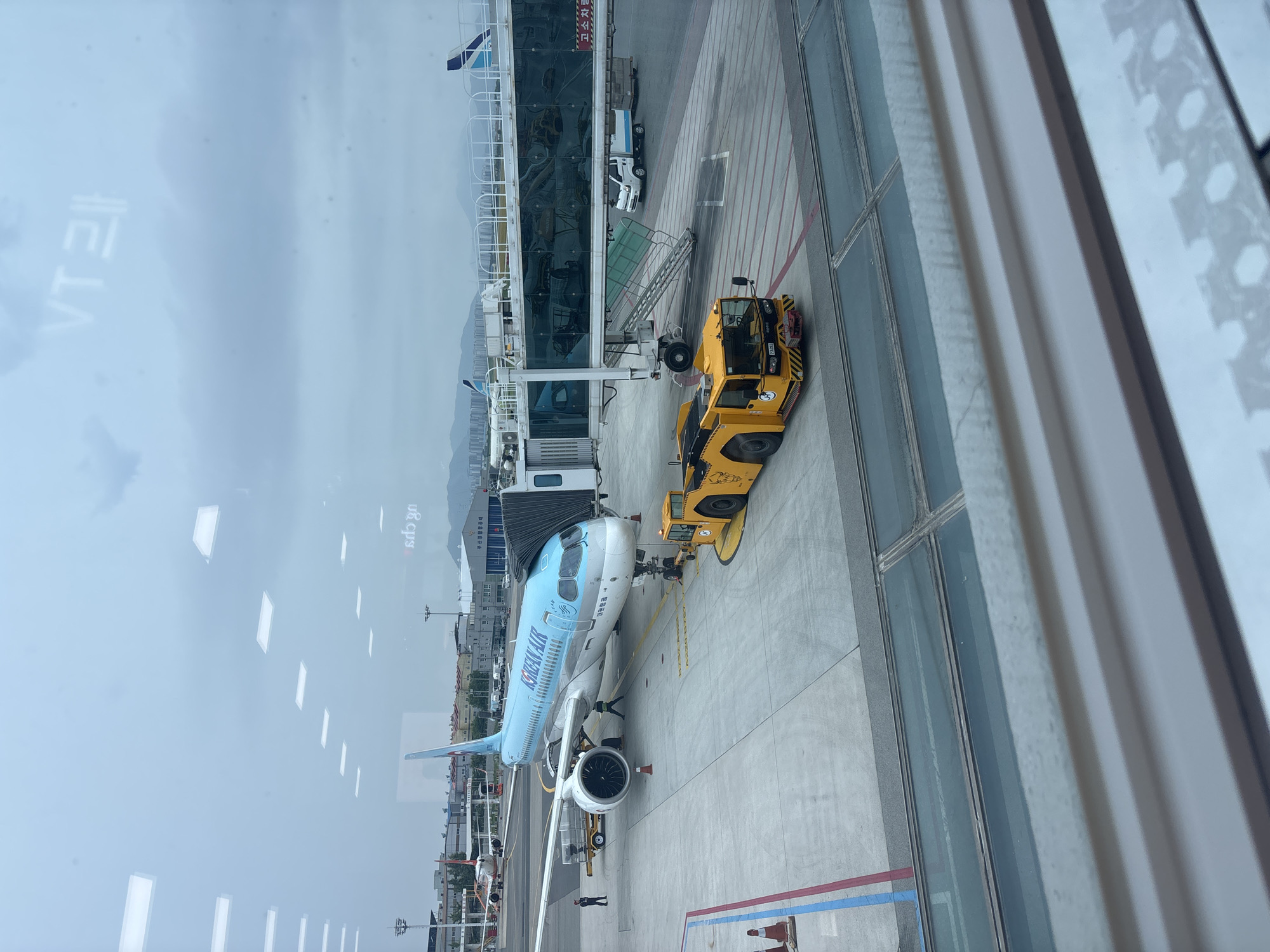 This image depicts Gimhae International Airport in Busan, South Korea. A Korean Air airplane is parked at Gate 7, connected to the terminal by a jet bridge. The aircraft, an Airbus A220-300, is painted in the airline's livery. A yellow aircraft tug is positioned near the nose of the plane, and several ground crew members are visible on the tarmac. In the background, other aircraft and airport buildings can be seen, along with a cloudy sky and distant mountains. The image appears to have been taken from inside the terminal, looking out through a window.