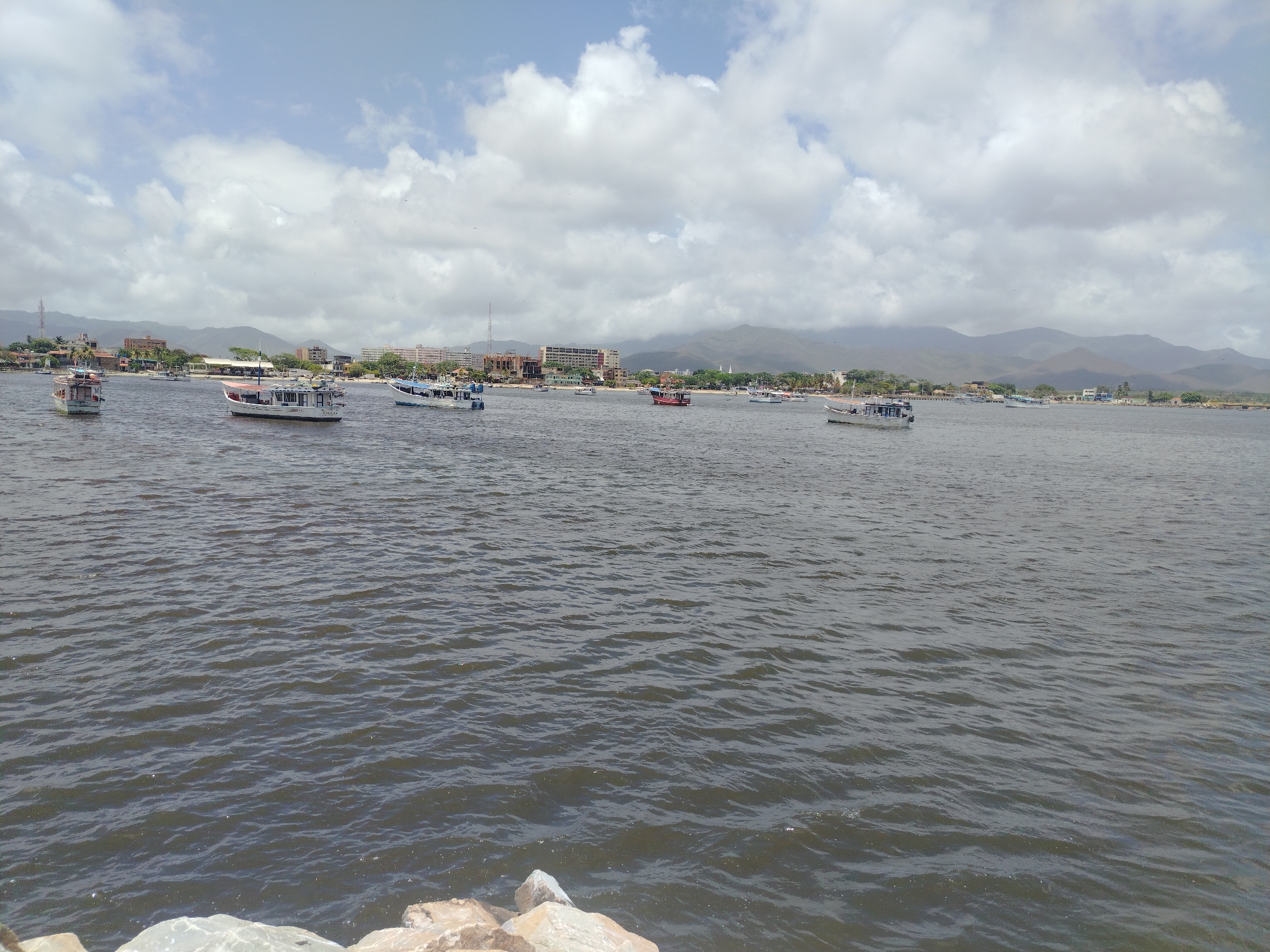 The image depicts a wide, tranquil body of water, likely a bay or gulf, in Juan Griego, Venezuela. The water is a dark, murky blue, with subtle ripples indicating a gentle breeze. Several boats of varying sizes are scattered across the water. In the background, a coastal town is visible, characterized by a line of buildings ranging from low-rise structures to taller apartment complexes and hotels. Hills and mountains form a dramatic backdrop to the town, with some appearing hazy due to distance or atmospheric conditions. The sky above is a bright blue, dotted with numerous fluffy white clouds. The foreground shows a few rocks and pebbles along the edge of the water, suggesting the vantage point is from a shoreline or a pier. The scene is calm and appears to be during daylight hours.