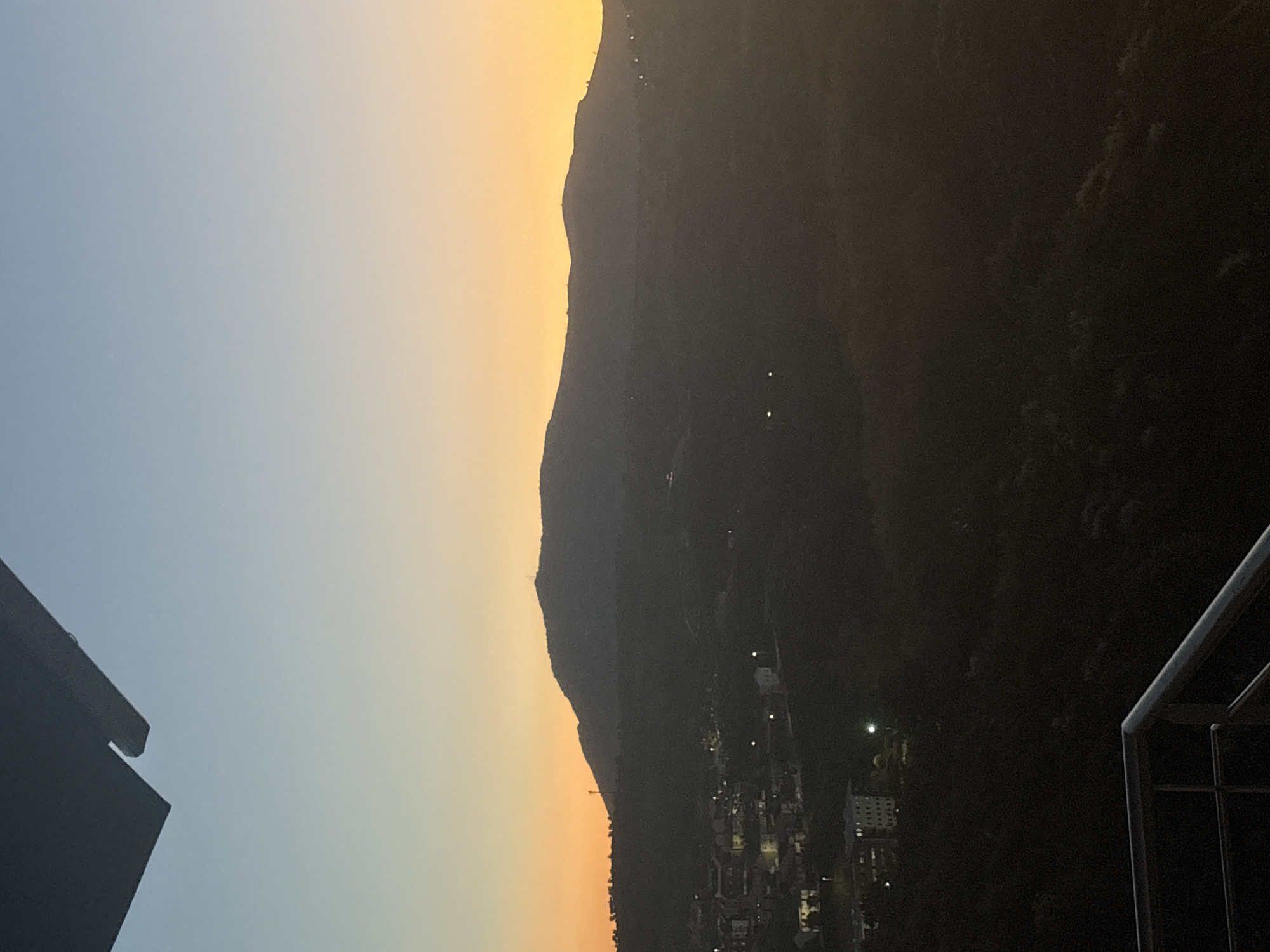 The image shows a dusk or twilight view with a silhouette of a mountain range against a gradient sky, transitioning from blue to orange. The slopes of the mountain seem to harbor a town or village, indicated by scattered points of light. There's part of a building in the foreground on the left and a railing on the right. The location could potentially be Mirandela, Portugal, given the hilly landscape and the presence of a settlement.