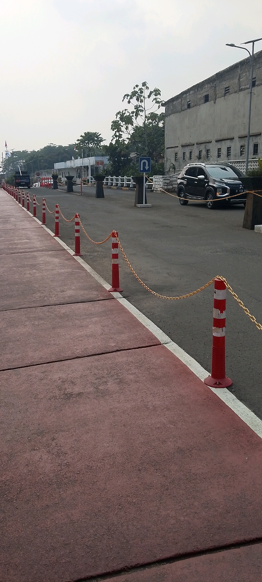 The image depicts an outdoor scene in Depok, Indonesia, likely near a government or administrative building, possibly the Depok City Hall complex. In the foreground, a red-colored pedestrian walkway is visible, segmented by expansion joints. A white line demarcates the edge of the walkway from the adjacent asphalt road. Along the road, a line of red and white striped stanchions connected by a gold chain are installed, serving as a barrier.
Further down the road, a dark-colored SUV is parked, with a license plate visible that reads "B 1708 DEF". In the background, a multi-story concrete building with visible windows and unfinished brickwork dominates the right side of the frame. Trees and greenery are scattered throughout the scene, with a U-turn traffic sign visible on the road. A lamppost stands near the building. A truck with a load bed is also visible on the left side of the image, further down the road. The sky is overcast, indicating a cloudy day. The overall impression is one of an urban infrastructure area, likely a public space or a service road adjacent to a significant facility.