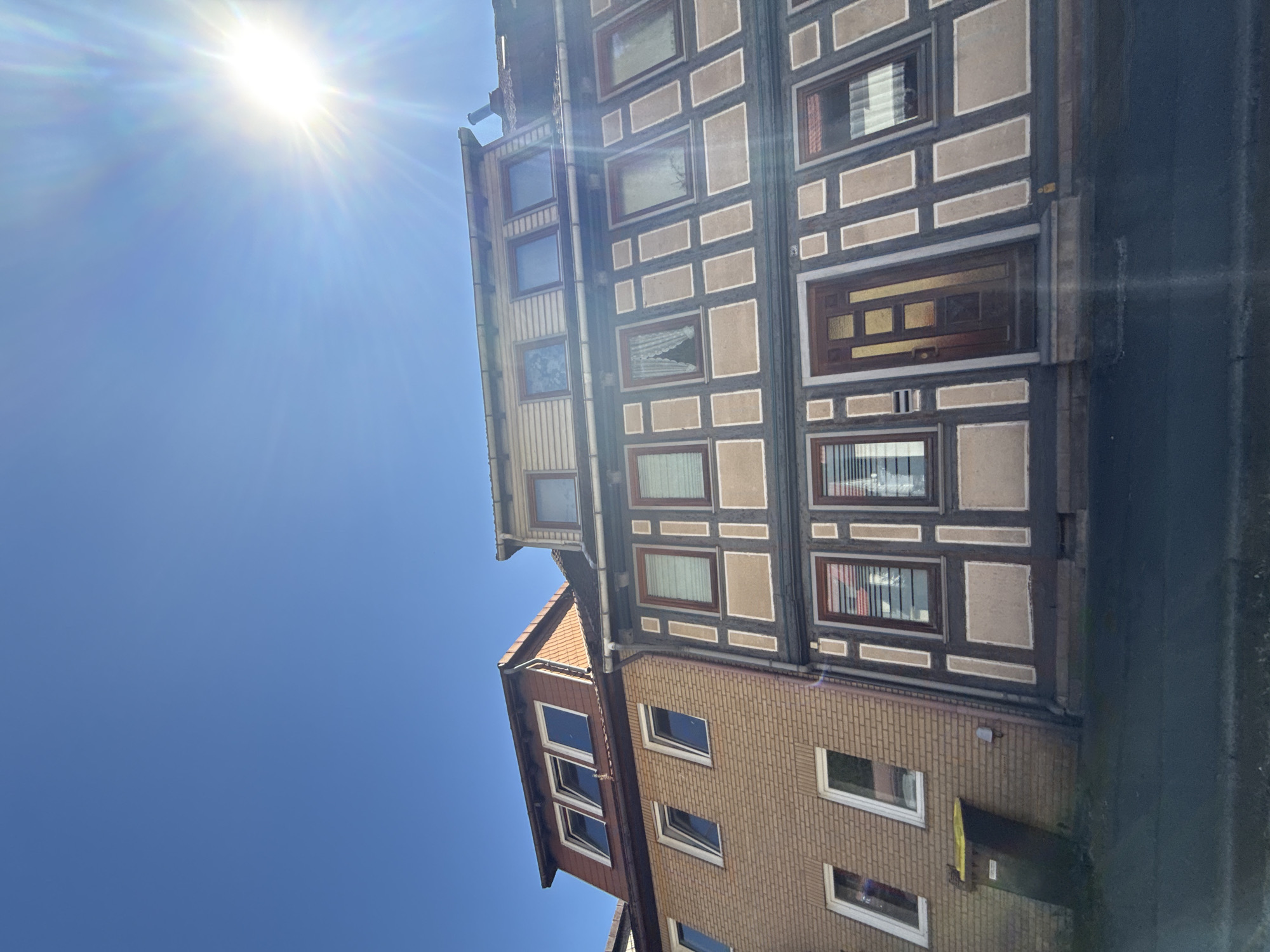 The image displays a close-up, low-angle view of two adjacent buildings under a clear blue sky with the sun shining brightly and causing lens flare. The building on the left is a modern structure with light brown brickwork and several windows. It has a red-tiled mansard roof with multiple dormer windows. The building on the right is an older, traditional half-timbered house with dark brown structural beams and lighter colored infill panels. This building features numerous windows with dark wooden frames, some with lace curtains. A dark wooden door is visible on the ground floor. A yellow trash bin is situated next to the brick building, and a small yellow marker is on the ground in front of the half-timbered house. The image appears to be taken on a street in the town of Bad Lauterberg im Harz, Germany, given the architectural style of the older building, which is characteristic of many buildings in such historic German towns. There are no people visible in the image.