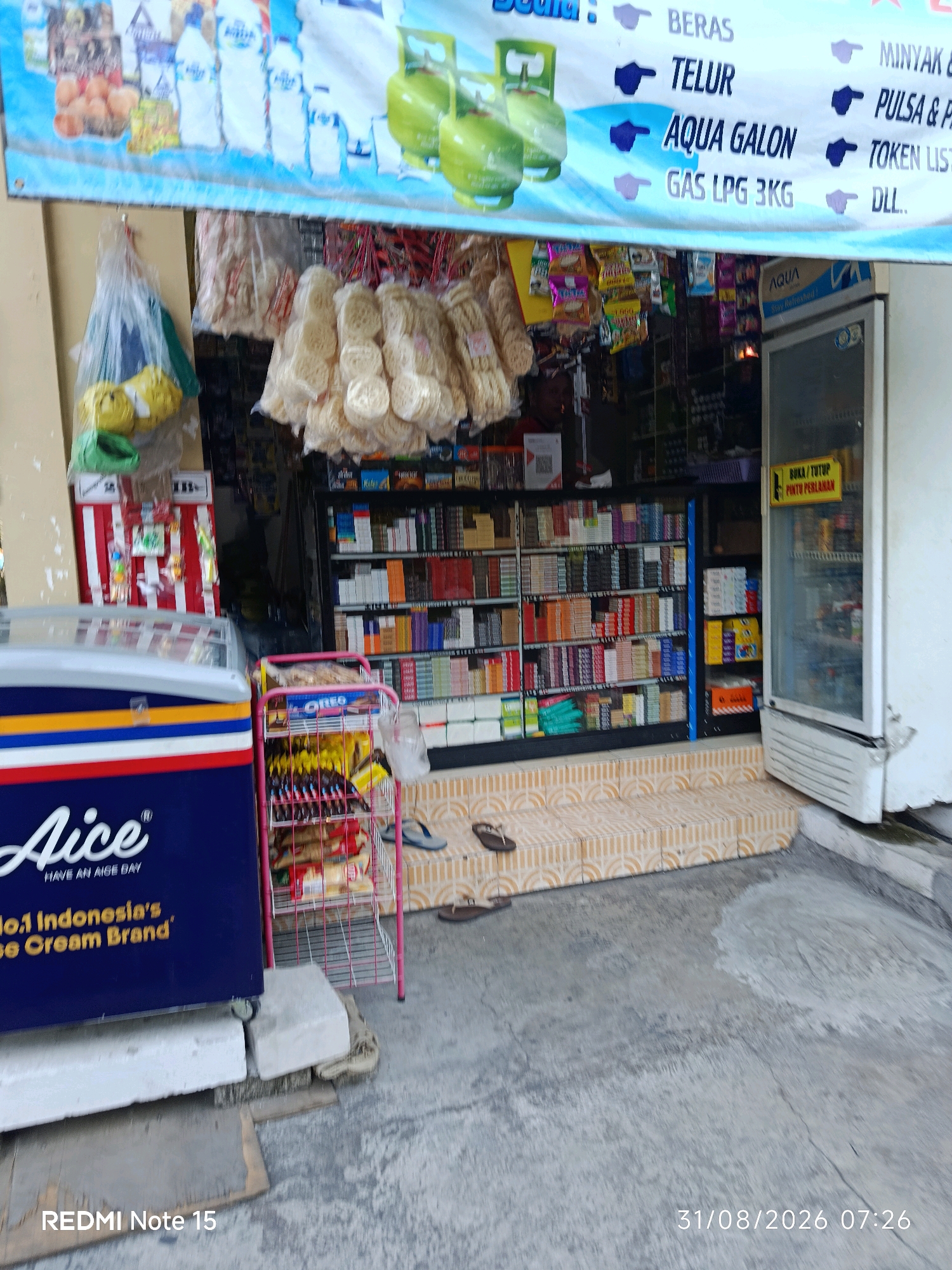 The image captures a street-side shop in Keboansikep, Indonesia, as indicated by the timestamp and the style of the storefront. In the foreground, a blue and white Aice ice cream freezer is prominent, displaying "No.1 Indonesia's Ice Cream Brand". To the right of the freezer is a pink metal display rack filled with various snacks and products, including a visible package of Oreo cookies. Behind these, shelves are stocked with numerous cigarette packs, and further back, a white refrigerator stands open, showcasing drinks and other items. A blue and white banner hangs above the storefront, listing products such as "BERAS", "TELUR", "AQUA GALON", and "GAS LPG 3KG", along with other services like "MINYAK", "PULSA & P", and "TOKEN LIST..". The shop appears to be a small convenience store or warung. The ground in front of the store is paved with concrete and tiles. Two pairs of flip-flops are visible on the tiled step leading up to the shop. The overall scene depicts a typical retail environment in a local Indonesian neighborhood.