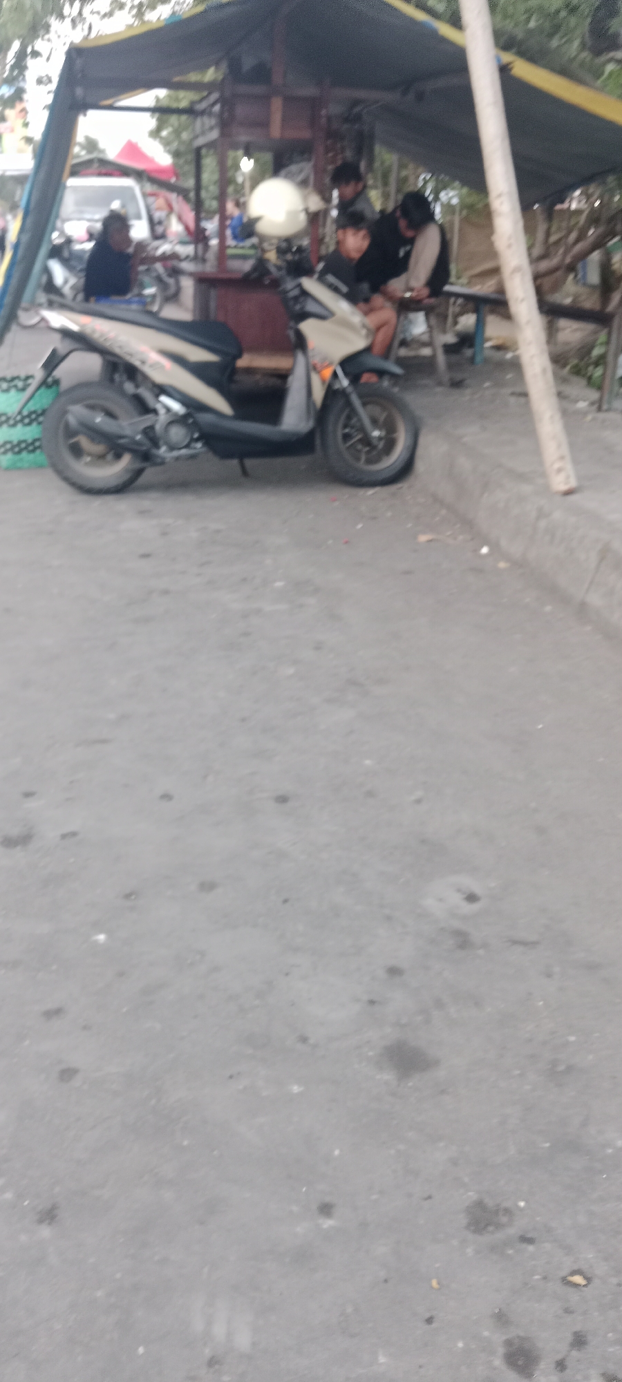 In Ngemplak, Indonesia, a tan and black scooter is parked on a concrete surface, its rear tire resting near a curb. Behind the scooter, a temporary shelter with a dark tarp roof is erected. Within the shelter, three young men are present. One is seated on a bench, leaning forward and looking down, while another stands behind him. A third individual sits on a stool, also looking down. A white helmet rests on the back of the scooter. The environment appears to be an outdoor setting with some vegetation visible in the background and other vehicles, including a white van, further in the distance. The overall scene suggests a casual gathering or stop at a roadside vendor.