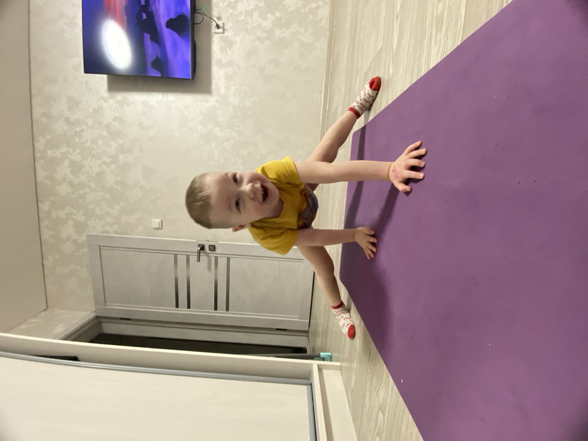 A young boy in a yellow t-shirt and Christmas-themed socks is crawling on a purple yoga mat in a room. The room features a TV displaying an image, textured wallpaper, a white door, and a white storage unit with an open compartment. The location is a domestic interior. There is no evidence to suggest it is in Sochi, Russia.