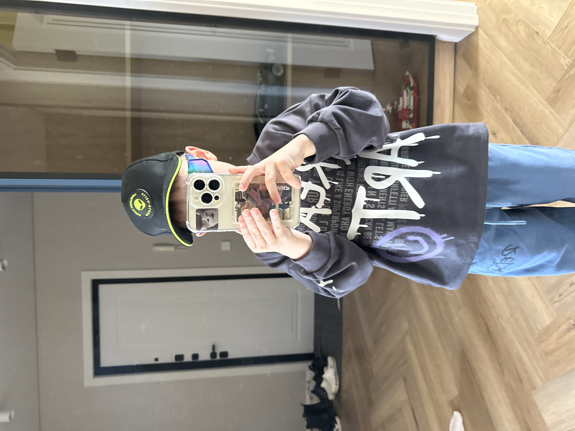 A child is standing indoors, holding a smartphone to take a photo or video of their reflection in a large glass surface. The child is wearing a black cap with a yellow design, a gray long-sleeve shirt with white text and a purple swirl design, and blue jeans. Behind the child is a glass panel. Behind the glass is a door. Below the door, there are multiple pairs of shoes. The location appears to be in Chaikovsky, Russia.