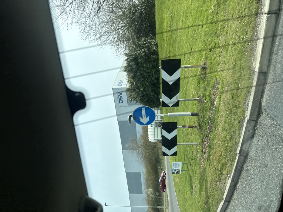 The image, taken from within a vehicle, captures a street scene at a roundabout in Thrapston, United Kingdom, specifically near a DSV logistics facility. The foreground shows the interior top edge of a vehicle's windshield, including a rearview mirror housing and a dashcam or sensor unit. Parallel lines, possibly defrosting elements, are reflected on the windshield.
The scene focuses on the central grassy mound of a roundabout, covered in short green grass with scattered small white flowers. Three traffic signs are centrally positioned on this mound: two rectangular black signs with white left-pointing chevrons, and one circular blue sign with a white left-pointing arrow mounted above them on a taller pole. To the left of these signs, a smaller white sign with black text, partially visible, displays "A C BOWNS" and contact details, likely an advertisement. Behind the signs, a white skip or trailer with a green lid is visible on the mound.
An asphalt road surface with a concrete kerb defines the edge of the roundabout. On the far side of the roundabout, a red hatchback car, possibly a Mazda model, is observed moving right-to-left. In the background, a large, light grey industrial building with the "DSV" logo displayed prominently multiple times on its facade is visible. The area around the building and the roundabout is bordered by various trees and bushes, some with bare branches and others with green foliage. A street light pole stands on the far left. The sky is clear and bright. No individuals are visible in the frame.