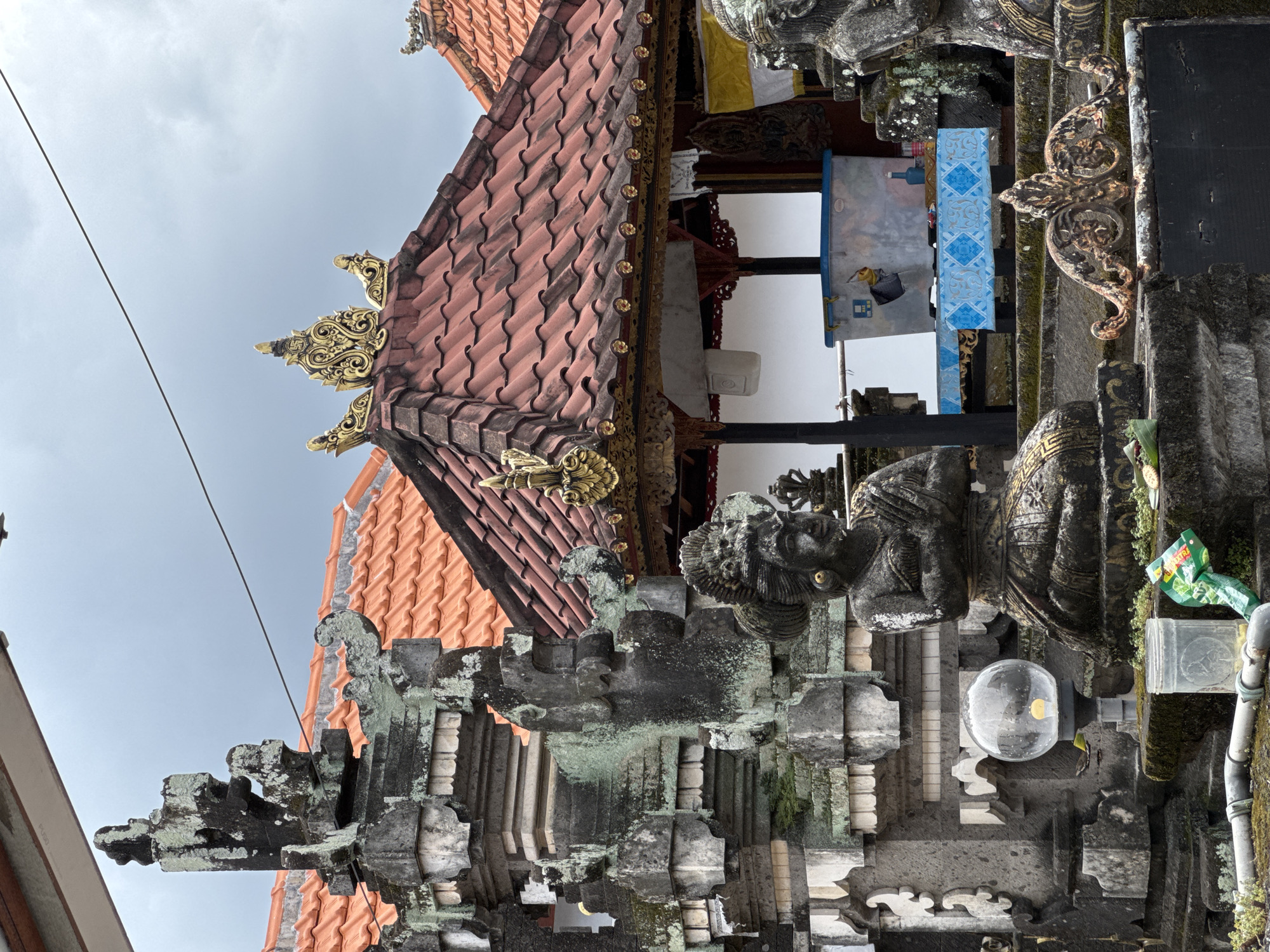 The image depicts a structure in North Kuta, Indonesia, featuring a terracotta tile roof with ornate gold trim. Below the roof is a white wall with a blue patterned cloth hanging on it. In the foreground, there is an aged stone structure adorned with a weathered stone statue and a clear round lamp. Moss grows on the stone, and there's a drink container attached to it. The sky above is overcast.