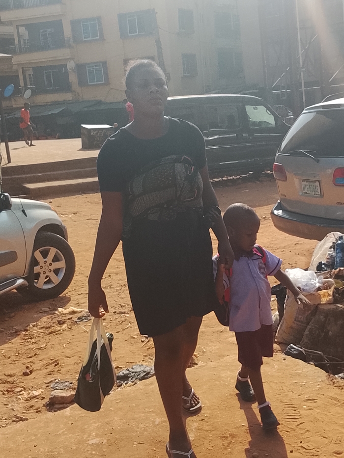 A woman walks with a child on a sunny day. She carries a black bag and wears a black dress and white sandals. The child wears a uniform with a backpack. They are walking on a dirt road near parked cars. A building and other people are visible in the background.