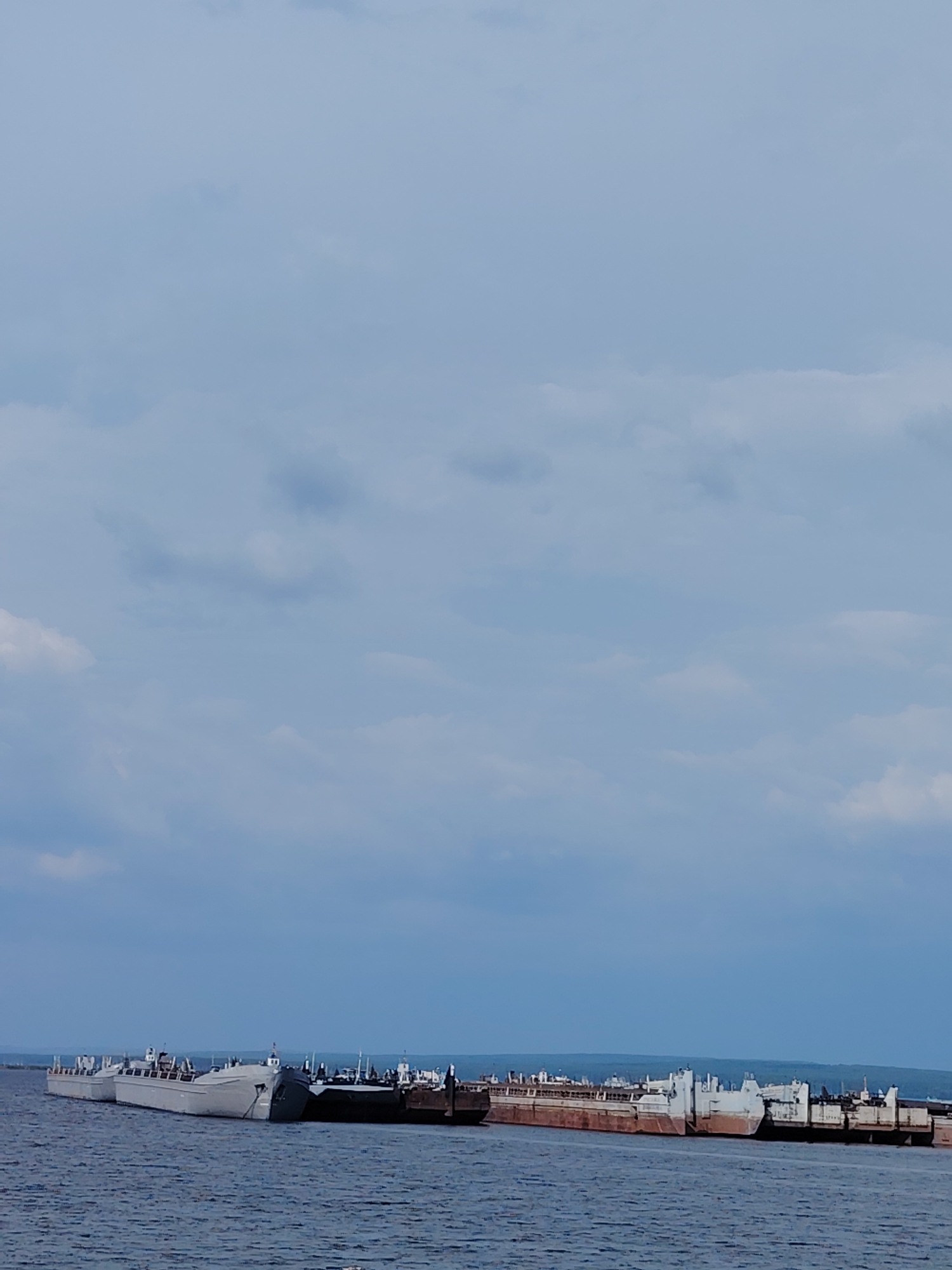 The image shows a view of the Kama River in Chaikovsky, Russia, with a fleet of barges moored in the water. The sky is overcast with soft grey clouds, and the water is calm and dark blue. The barges are lined up along the shore, with some appearing to be loaded with cargo. The scene appears to be one of industrial activity, with the barges likely used for transporting goods along the river. The horizon is distant and blurred, with a faint line of trees or land visible in the background.