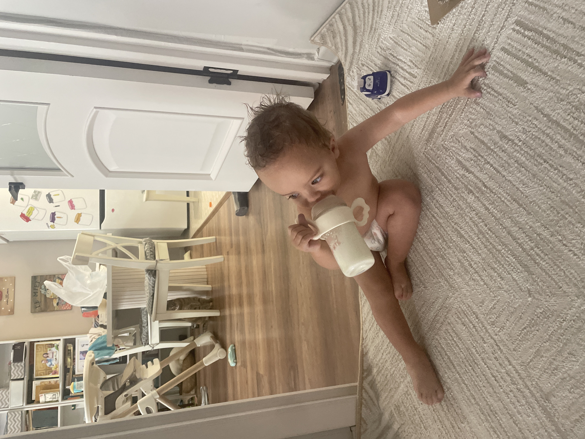A young child is sitting on a textured rug, drinking from a bottle. The child is wearing a diaper and appears to be in a home environment. In the background, a white door with panels is visible, along with a wooden floor. There are also glimpses of furniture, including chairs, a high chair, and bookshelves, suggesting a living area or nursery. A small blue and white toy is on the rug near the child. The lighting suggests it might be daytime. The overall impression is a candid moment of a child being cared for.
