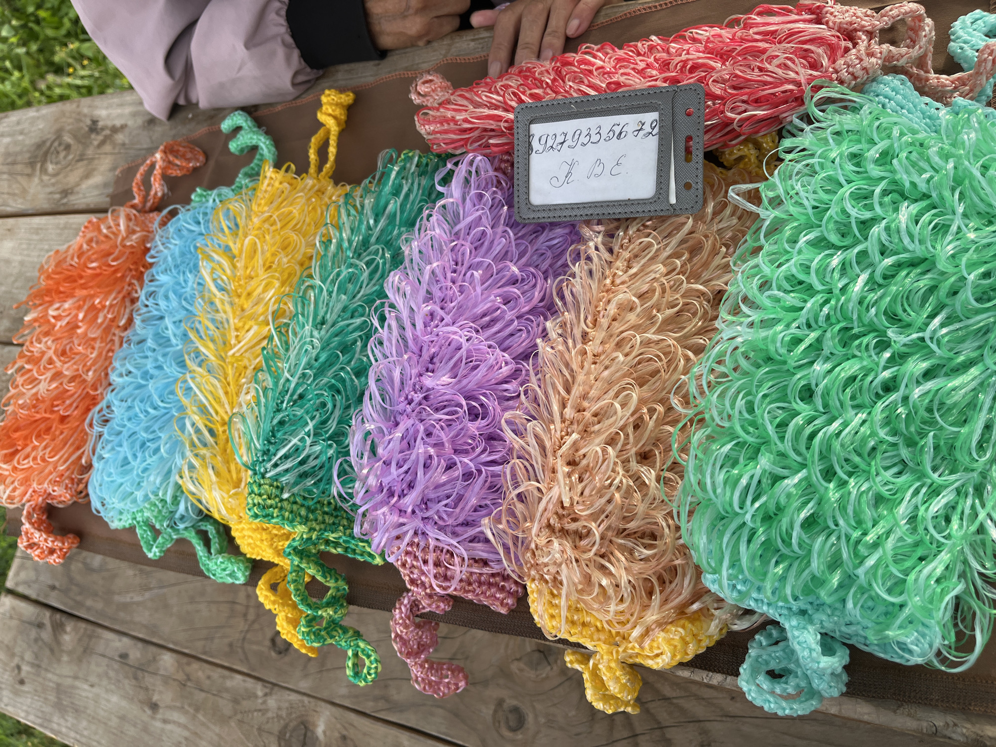 A collection of brightly colored, crocheted mesh items, likely bath poufs or scrubbers, are laid out on a weathered wooden surface. The items are arranged in a diagonal line from the top left to the bottom right of the frame, with colors including orange, light blue, yellow, green, purple, light peach, and red. A gray luggage tag with handwritten markings is placed on top of one of the items. The background shows a hint of green foliage and a portion of a person wearing a light purple jacket, suggesting an outdoor setting. The image was taken in the city of Blagoveshchensk, Russia.