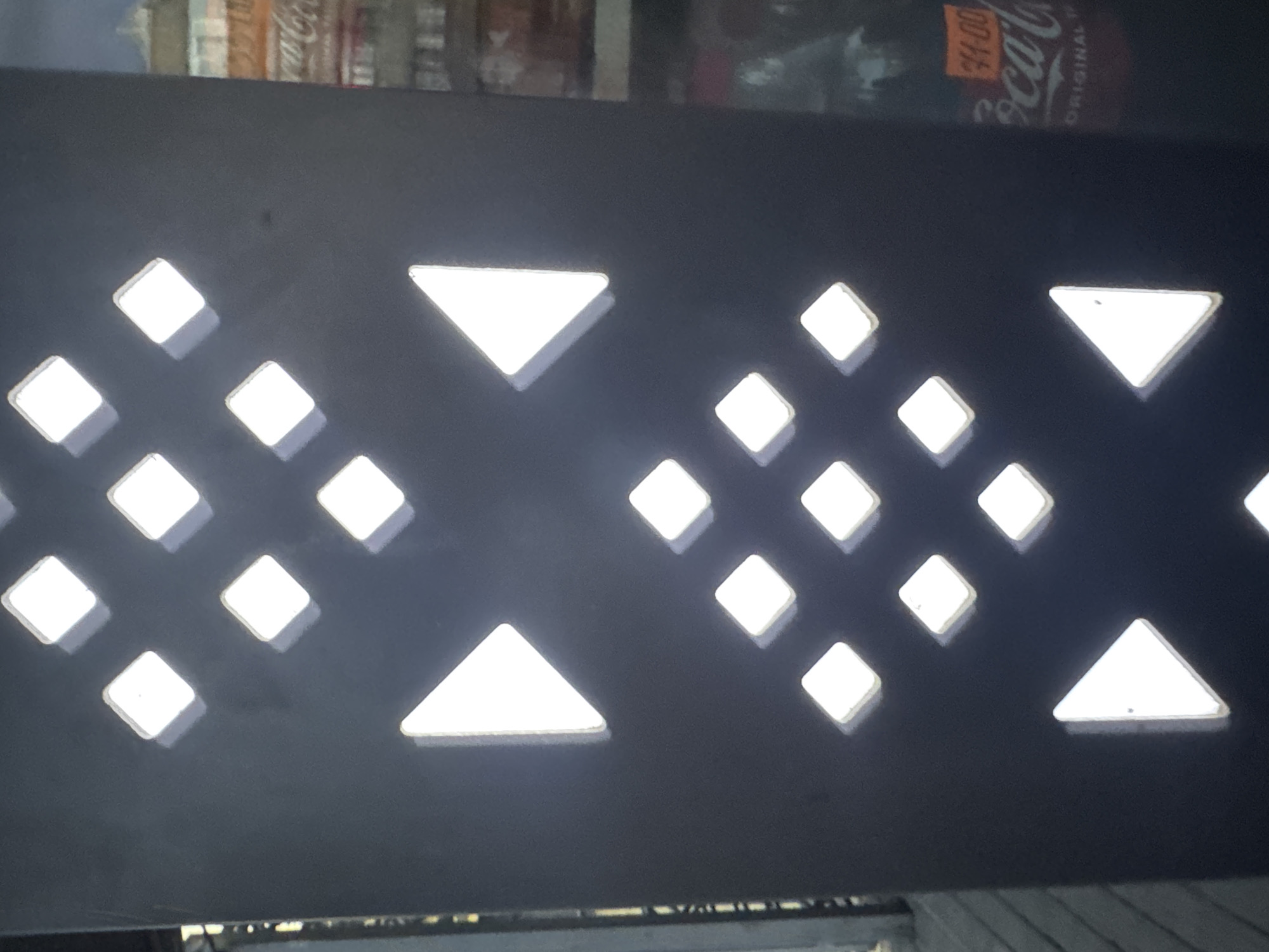 The image shows a close-up view of a dark, vertical structure with illuminated diamond and triangle shapes cut out of it. The diamonds are arranged in a grid pattern, with some rows offset from others. The triangles appear to be pointing left and right, arranged in a symmetrical pattern. In the background, a blurry reflection shows a store or building with a Coca-Cola advertisement. The scene appears to be outdoors, possibly on a street or walkway, given the presence of a wooden surface to the left. The specific location in Kremenchuk, Ukraine, cannot be determined from the image alone.