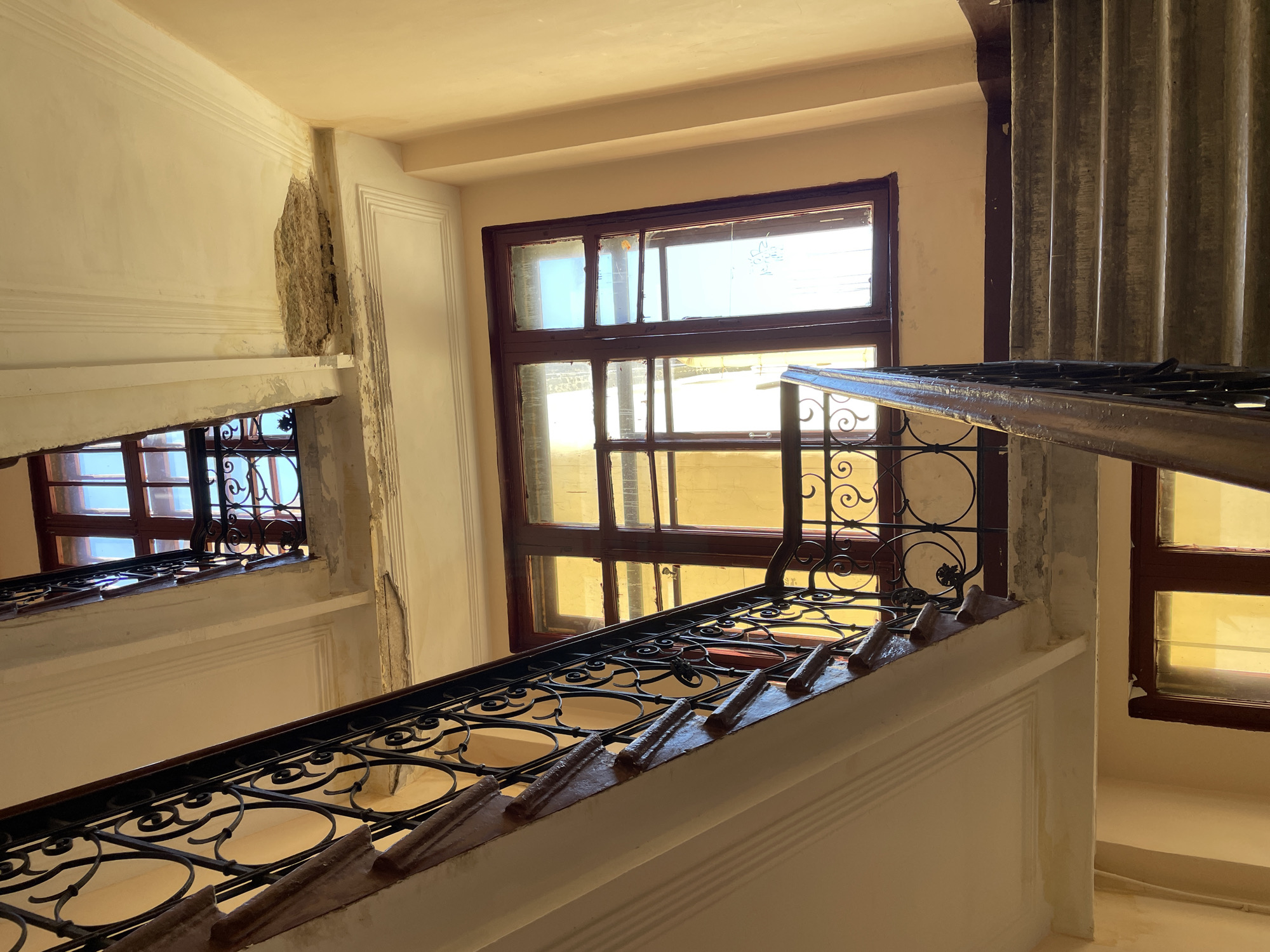 The media file depicts a view looking down a staircase with ornate black metal railings and cream-colored walls. The staircase has a wooden handrail. Multiple windows are visible on the left and right sides of the staircase. The windows have dark wooden frames and are divided into multiple panes. The glass in some panes appears frosted or dirty, with light filtering through, suggesting it is daytime.
On the left side, a window is visible with decorative metal grilles. Behind this window, another window with similar grilles can be seen further down the staircase. The wall to the left of the staircase shows some damage, with plaster broken away in one section, revealing the rough material beneath.
On the right side of the staircase, a large window with dark wooden frames is prominent. The glass in this window is tinted yellow, indicating light from outside. Further down, another window with a similar yellow tint is partially visible.
At the top of the frame, on the right, a corrugated metal structure is present, adding an industrial element to the otherwise classical architectural style of the staircase. The overall impression is that of an older building, possibly an apartment block or a historic structure. The lighting suggests it is either late morning or early afternoon, with sunlight casting shadows and highlighting the textures of the walls and railings. No people are visible in the frame. No text is present. The scene appears to be located in Saint Petersburg, Russia, as indicated by the location context.