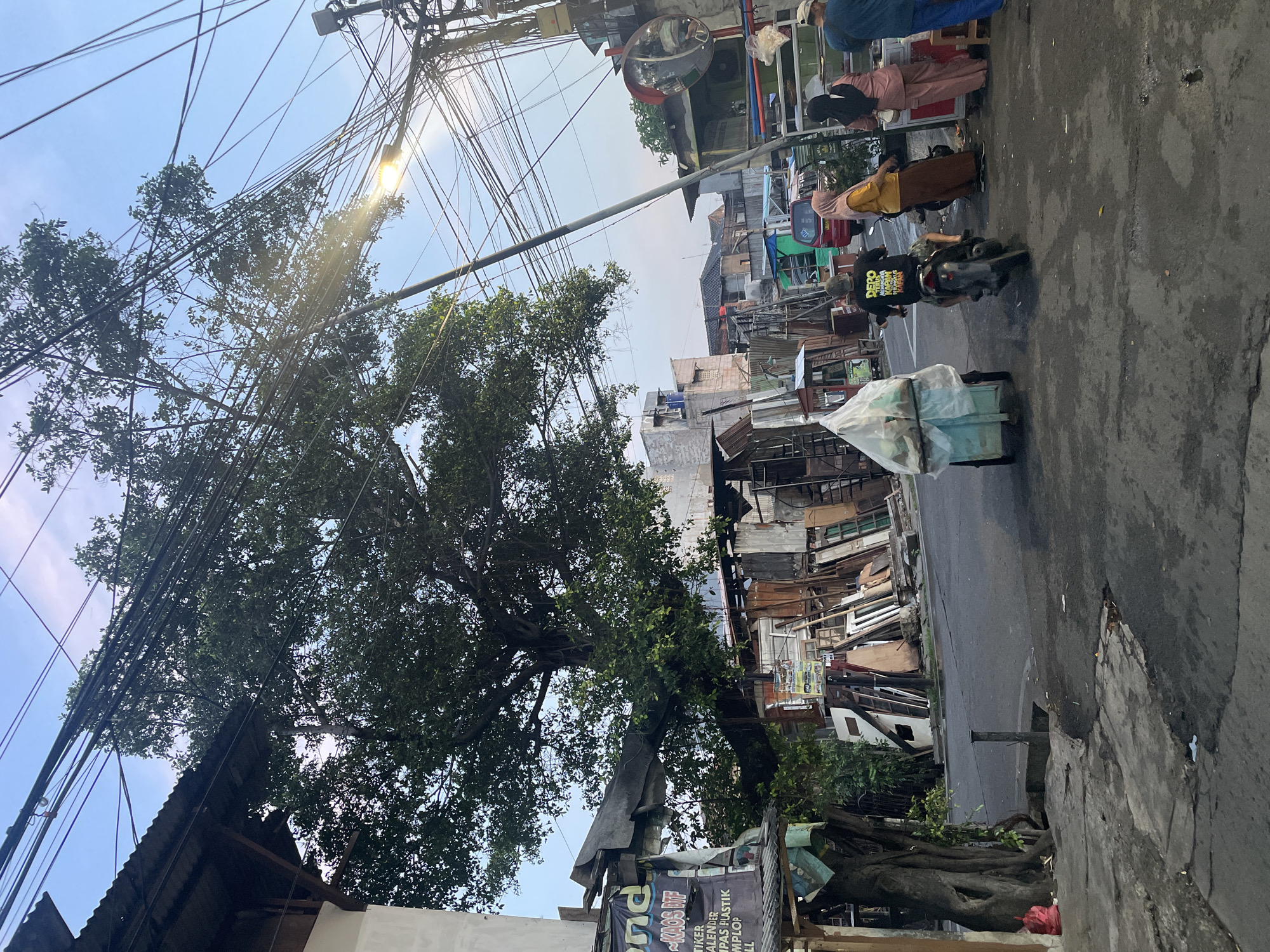The image, taken in North Jakarta, Indonesia, portrays a typical urban street scene. Overhead, a dense web of electrical wires obscures the sky, intertwining with a large tree. A convex mirror is mounted high on a utility pole. Along the right side of the frame, a street vendor stands with several customers. A motorcyclist passes them, pulling a small cart covered with a white tarp. The street is lined with dilapidated buildings, some with open windows revealing stored materials. A sign is partially visible in the foreground, featuring a list of items. The street itself is paved but shows signs of wear.