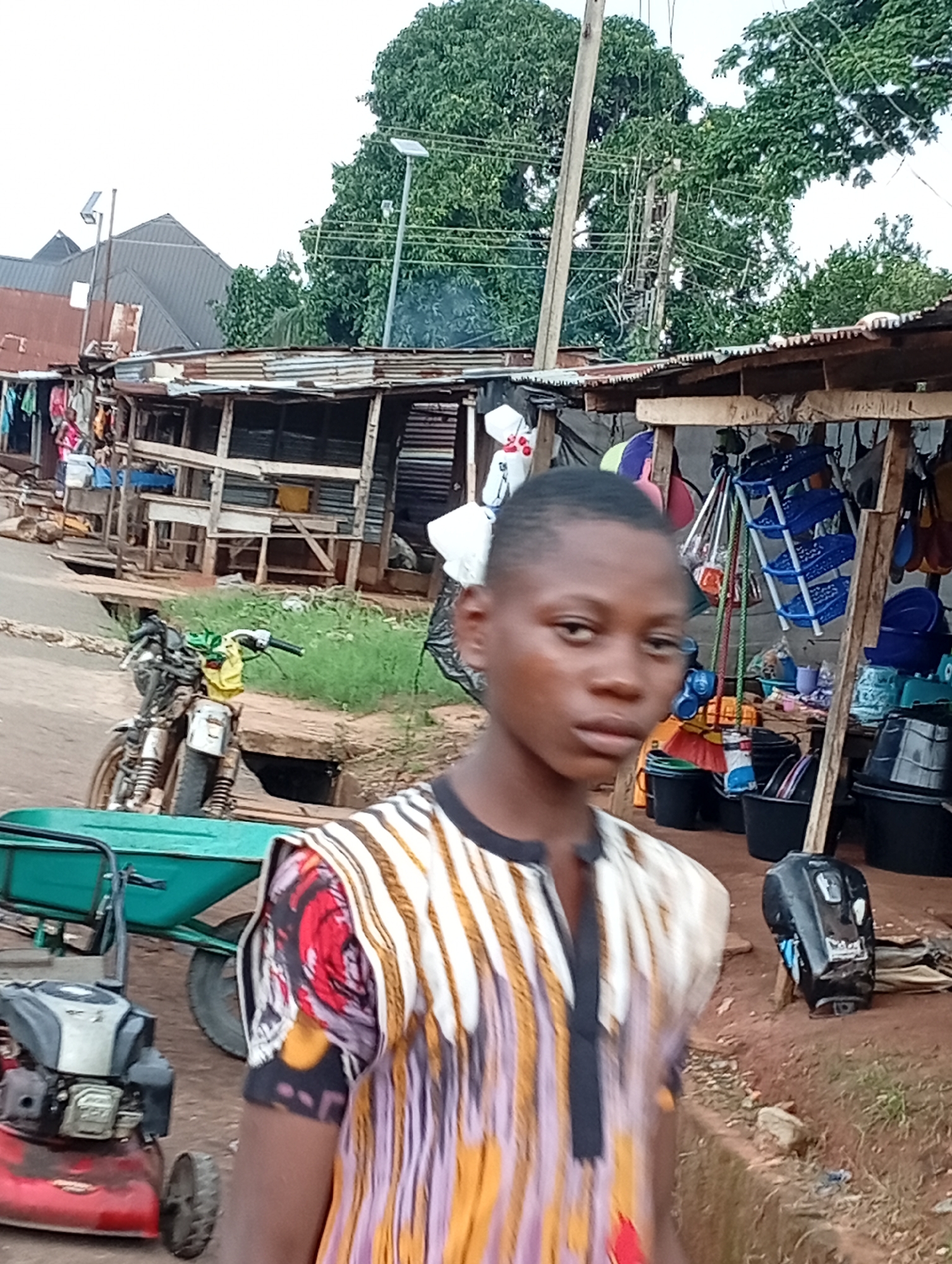 The image depicts a street scene in Nsugbe, Nigeria. In the foreground, a young person stands close to the camera, wearing a colorful, patterned shirt. Their gaze is directed forward. Behind them, a motorcycle with a yellow cloth tied to its handlebars is parked on a dirt road. To the left, a red lawnmower sits on the ground. Further back, a row of market stalls with corrugated iron roofs is visible, displaying various items, including buckets, baskets, and clothing. The stalls appear to be made of wood and corrugated metal. Trees and power lines are visible in the background, suggesting an outdoor, semi-urban environment.