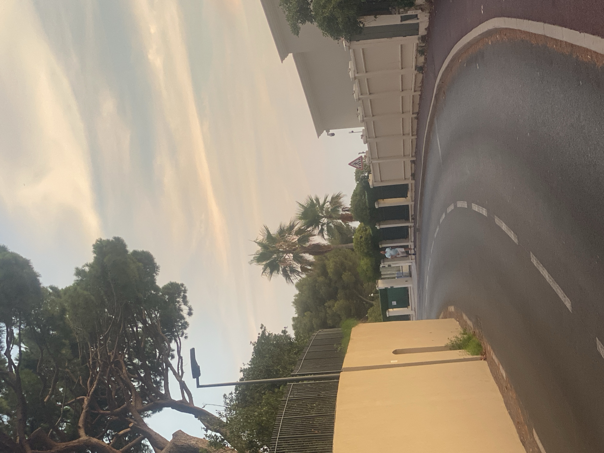The image shows a street scene in Antibes, France, during what appears to be late afternoon or early evening. A curved asphalt road occupies the foreground, with a white dashed line marking the lane. A lighter colored sidewalk is visible on the right side of the road. On the left side of the road, a tall, light-yellow wall runs along the street, topped with a dark metal fence. A large pine tree with dense foliage extends into the upper left corner of the frame.
Further down the road, on the right side, a man in a light blue polo shirt and shorts is walking away from the camera. He is in front of a white decorative fence that encloses a property. Beyond the fence, parts of buildings and more greenery, including palm trees, are visible. A traffic sign with a red border and a white triangle inside is also present near the fence. The sky above is filled with soft, pastel-colored clouds, suggesting a sunset or sunrise. The overall impression is one of a quiet residential street.