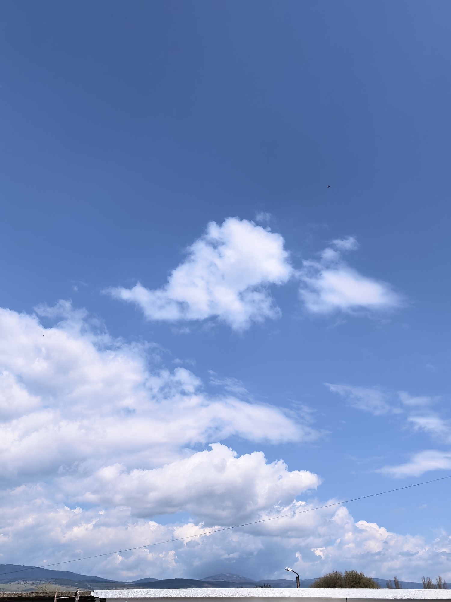 The image shows a daytime scene with a clear blue sky partially covered by white clouds. A small, dark object, possibly a bird or insect, is visible in the upper center of the frame. In the bottom portion of the image, a horizontal white structure is visible, behind which lies a landscape with trees and mountains. A lamppost extends upwards from behind the structure. A thin electrical wire stretches horizontally across the lower portion of the sky. The location is Unknown, Unknown.