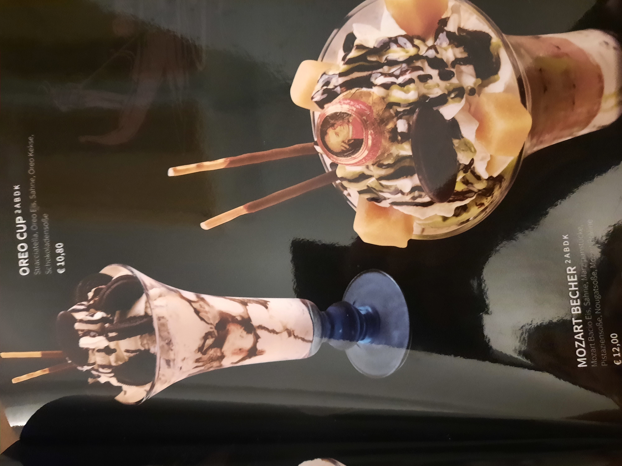 The image displays a menu from a restaurant, featuring two dessert items. On the left is an "OREO CUP" which costs €10.80. It is described as containing Stracciatella, Oreo Ice Cream, Cream, and Chocolate Sauce. The dessert is presented in a tall, stemmed glass, layered with ice cream and chocolate syrup, topped with whipped cream and Oreo cookies, and garnished with two chocolate-dipped biscuit sticks. To the right, a "MOZART BECHER" is shown, priced at €12.00. Its description includes Mozart Bacio Ice Cream, Cream, Marzipan pieces, Pistachio Sauce, and Nougat Sauce. This dessert is also served in a glass and is adorned with whipped cream, chocolate drizzle, fruit pieces, and two biscuit sticks, with a circular garnish featuring a portrait of a historical figure, possibly Mozart. The menu is set against a dark, reflective background, suggesting an indoor setting likely a restaurant or cafe. There is no discernible information about the specific location in Rosenheim, Germany, within the provided image.