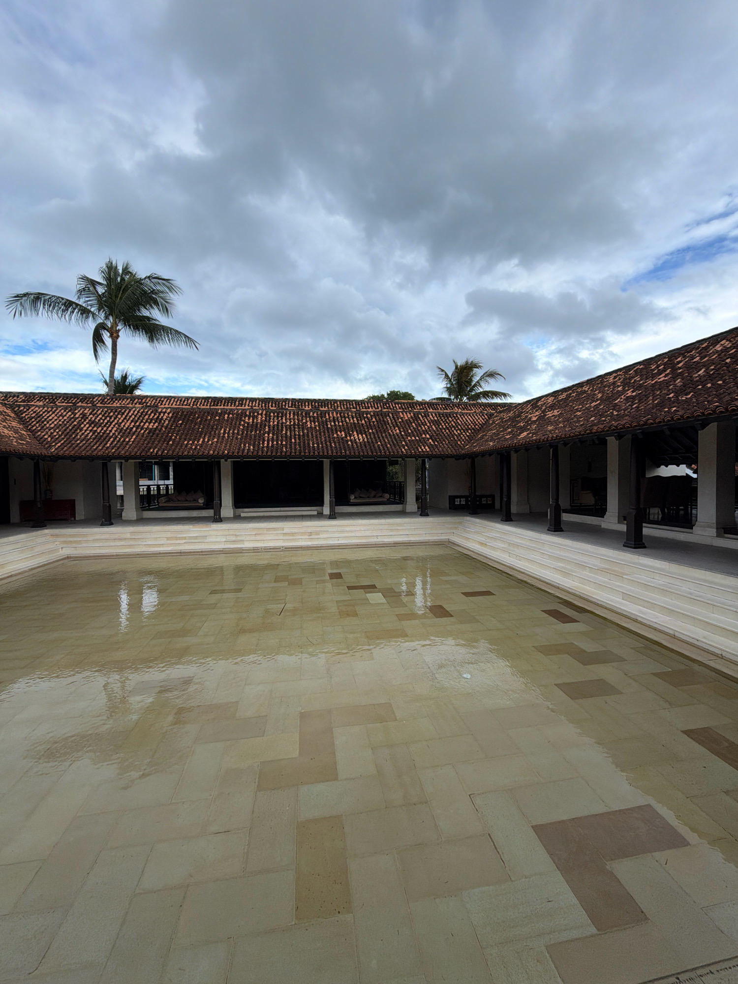 The image shows an outdoor courtyard with a shallow, wet tiled pool in the foreground. The pool's tiled floor is a mosaic of light beige and darker reddish-brown rectangular tiles laid in a parquet pattern. Water reflects the overcast sky and the covered walkway surrounding the courtyard. The walkway has a red-tiled roof supported by dark columns. Beyond the walkway, interior spaces with dark furnishings and large windows are visible. Palm trees stand behind the structure, silhouetted against a cloudy sky. The scene suggests a tranquil, possibly resort-like setting in Ko Samui District, Thailand, given the architectural style and vegetation. There are no people or animals visible in the image.