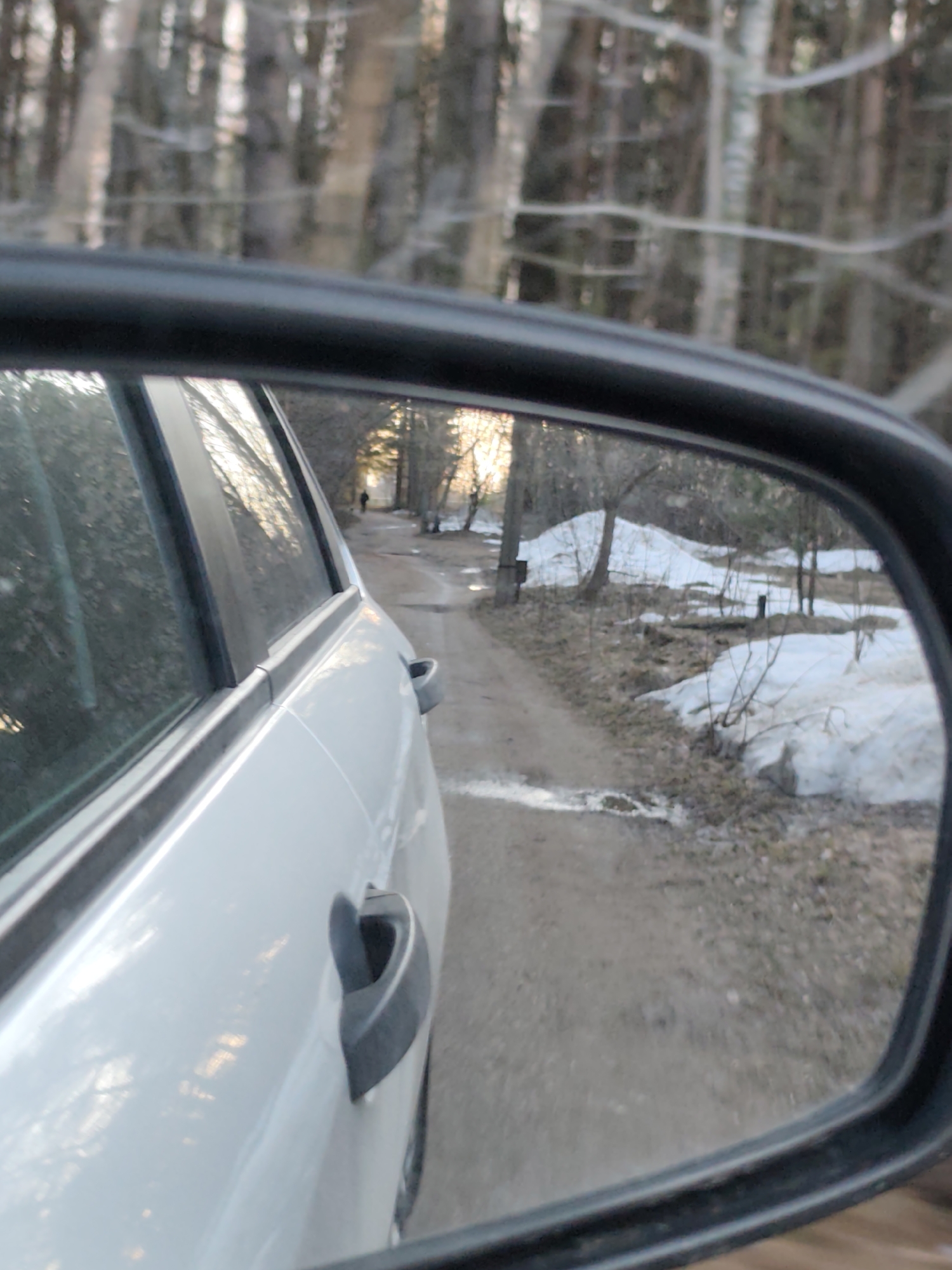 The image shows the side mirror view of a white car on a dirt road in a wooded area. The mirror reflects a dirt road with melting snow on the sides, partially covered by dried vegetation. A blurred figure is walking away on the road in the distance. The surrounding area has bare trees and vegetation. The foreground shows the side of the white car, including the door and handle. The location is presumed to be in a wooded area near Chaikovsky, Russia, based on prior information.