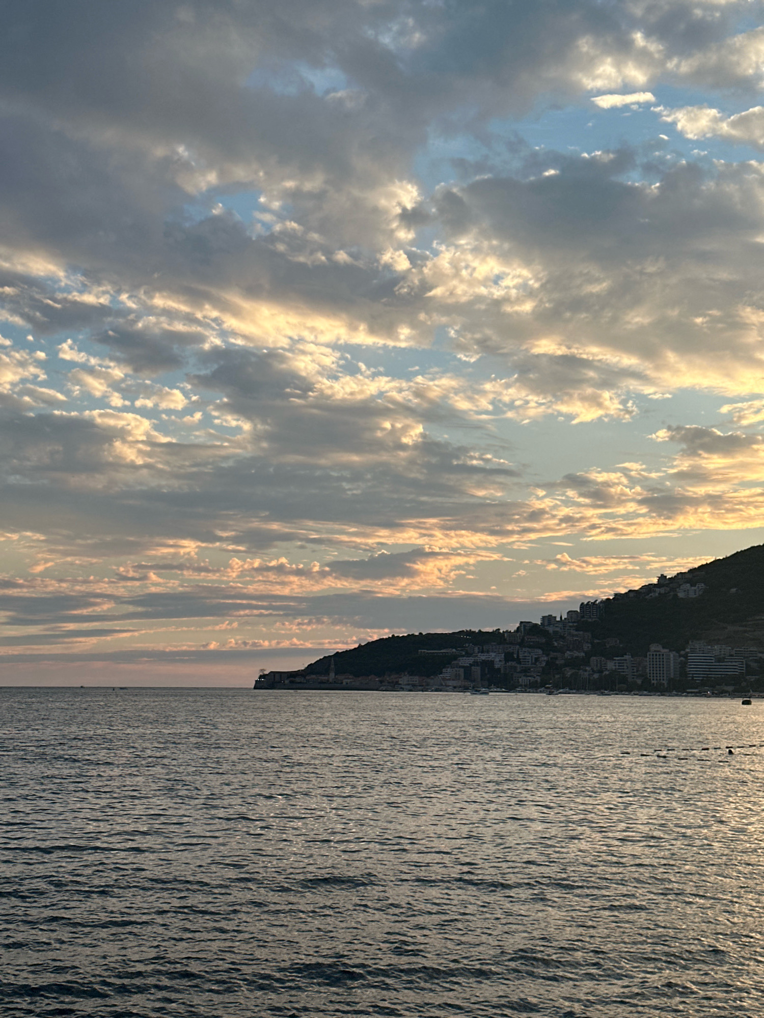 A wide shot shows the Adriatic Sea with a town built on a hillside in the background, under a dramatic cloudy sky. The sea is dark blue and rippled, with sunlight reflecting off the surface on the right side. The town, likely Budva, Montenegro, is built on a steep, tree-covered hill and features numerous buildings, some of which are white high-rises, clustered together. The hillside slopes down towards the water. The sky is filled with a mix of dark and light clouds, with patches of blue sky visible. Some clouds are illuminated by the setting sun, casting a warm, golden glow. There are no people clearly visible in the image, though there might be distant boats or figures on the water. The overall impression is of a scenic coastal town at dusk.