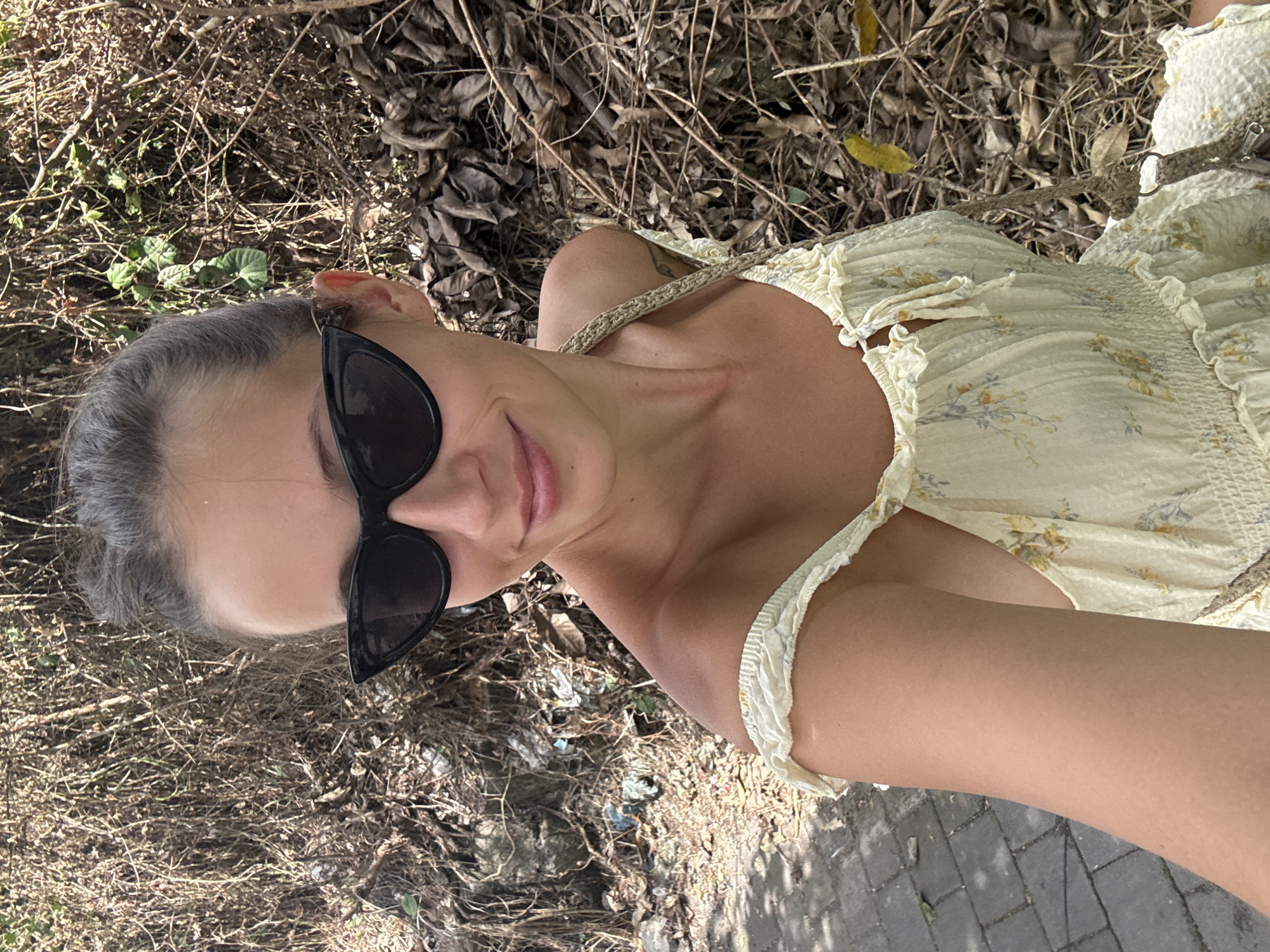 A woman wearing sunglasses and a light yellow floral dress is positioned in an outdoor setting. The background consists of dry twigs and foliage, with a section of stone pavement visible on the lower right. The location is not specified in the information provided.